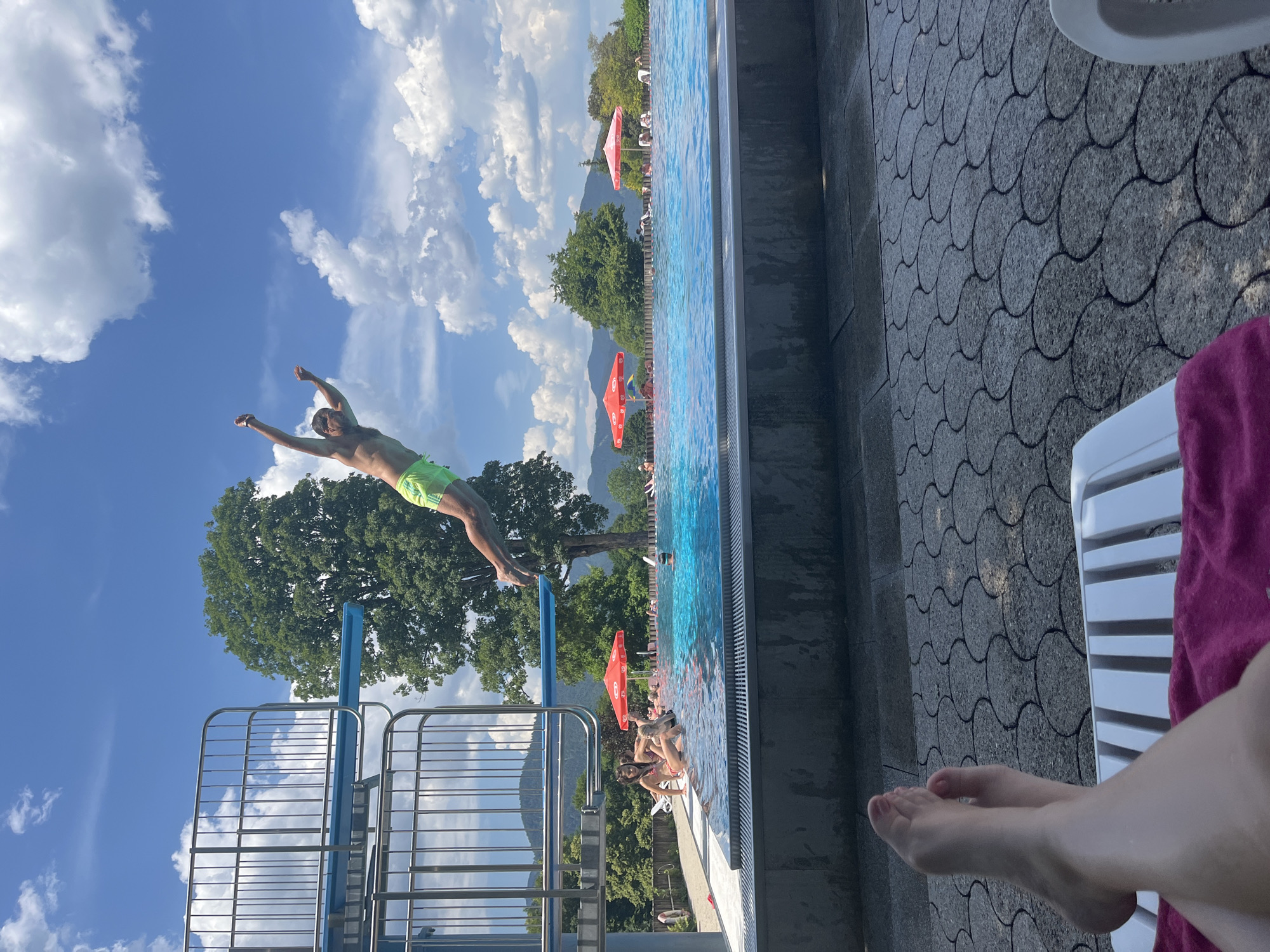 A young man in neon green swim trunks is captured mid-air as he dives from a diving board into a large outdoor swimming pool on a sunny day. He is in a horizontal position, with his arms extended upwards and outwards, his body angled downwards towards the water. The diving board is elevated, with metal railings on either side. The pool is a brilliant blue, with several people swimming and relaxing by the edge. In the background, lush green trees and rolling hills form a scenic backdrop under a bright blue sky dotted with fluffy white clouds. The foreground shows a person's legs and feet resting on a white lounge chair, with a pink towel draped over it, suggesting a relaxed atmosphere. The scene is set at a public swimming pool, possibly in the vicinity of Oberammergau, Germany, given the mountainous landscape and the architecture of the diving platform.