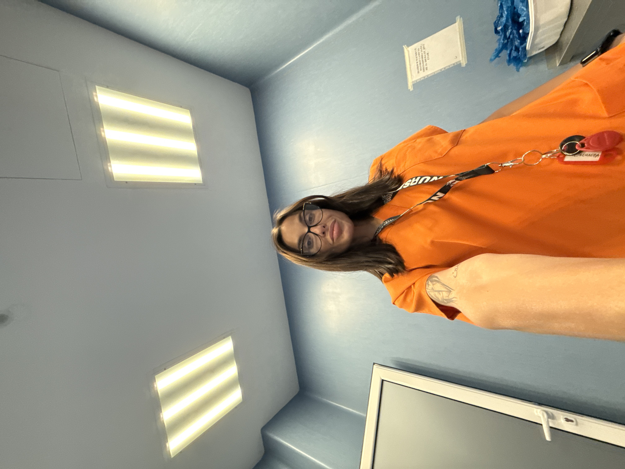 A low-angle, selfie-style shot features a woman with dark hair and glasses wearing an orange scrub top and a lanyard with keys and ID badges. She is standing in a room with light blue walls and a white ceiling with two rectangular fluorescent light fixtures. To the left, a white door with a silver handle is visible. On the right wall, a document is taped, and a white plastic container filled with blue plastic bags sits on a surface. A tattoo is visible on the woman's left arm. The setting appears to be a medical or institutional environment.