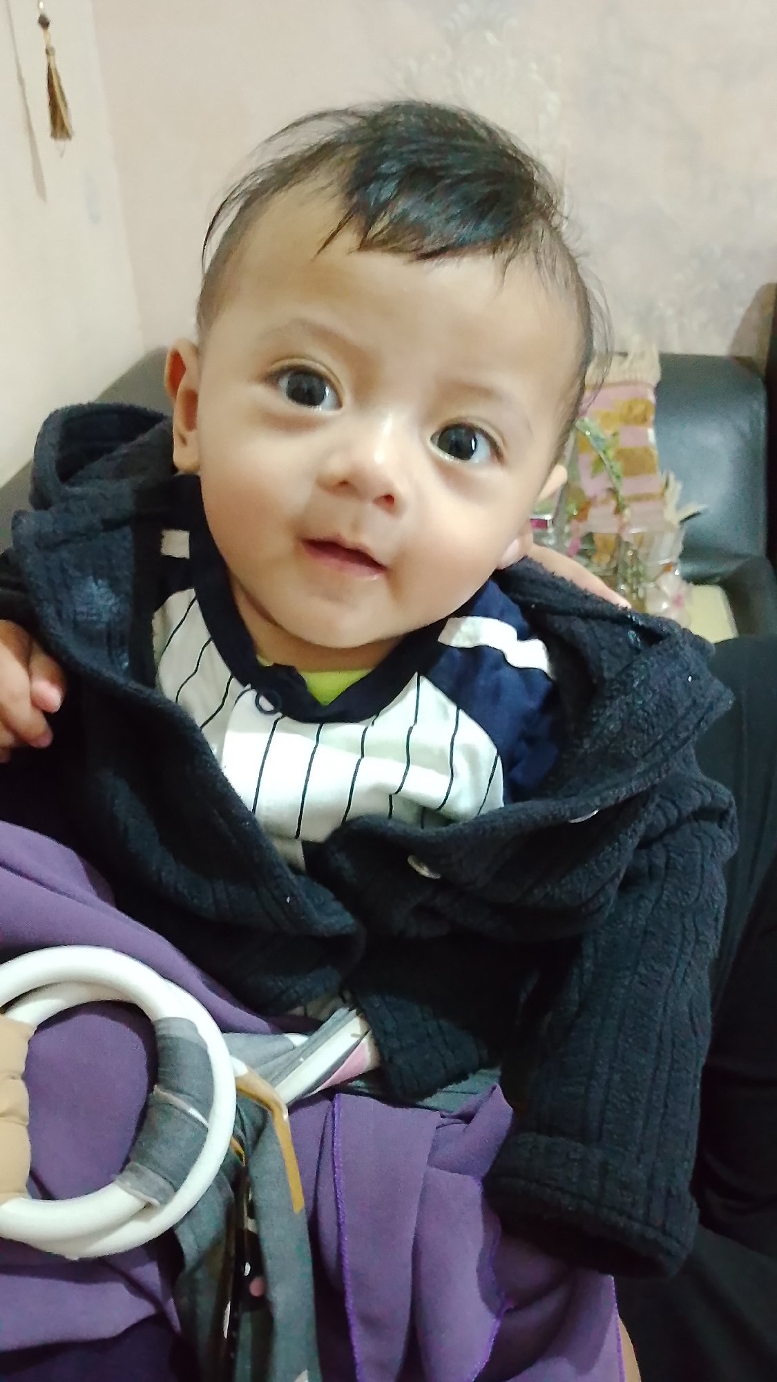 A close-up shot focuses on a young baby, likely an infant, looking directly at the camera. The baby has dark hair, large dark eyes, and fair skin. They are wearing a striped shirt with blue and white stripes and a lime green undershirt. A black hooded jacket is draped loosely around the baby, partially obscuring their body. The baby is being held, with a purple cloth visible beneath them. A white ring with grey and tan accents, possibly part of a toy or accessory, is in the lower left portion of the frame. The background is softly blurred, with hints of a pale wall and a dark piece of furniture, suggesting an indoor setting. The lighting appears to be natural or from a soft artificial source, illuminating the baby's face. The location is identified as Bojongsoang, Indonesia.
