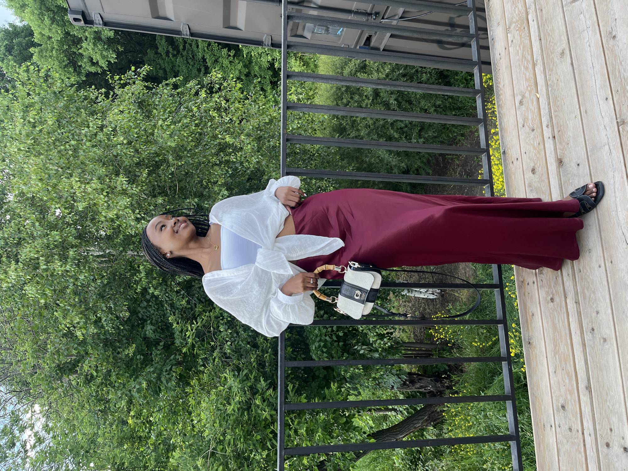 A young Black woman with braided hair stands on a wooden deck, leaning against a black metal railing. She wears a white, tied-front blouse over a white bandeau top and a long, burgundy satin skirt. A small, black and cream handbag with a bamboo handle hangs from her left shoulder. She is looking to her right with a slight smile. Behind her, lush green trees form a dense backdrop. To her right, a portion of a large, dark metal shipping container is visible. The deck has a light wood grain and is surrounded by a black railing. Below the railing, dense green foliage and scattered yellow flowers are visible.