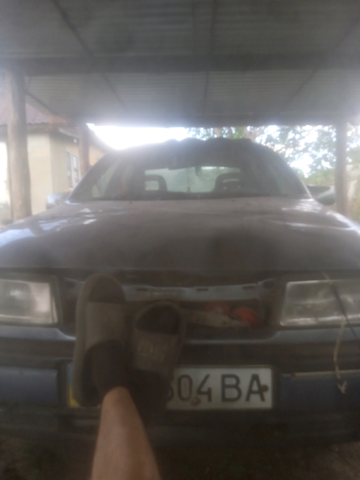 An overexposed, low-resolution, horizontal, medium shot photograph shows the front of a dark-colored car parked under a corrugated metal roof supported by wooden beams. The car appears to be an older model, possibly a sedan or hatchback, with visible headlights and a front grille area. A license plate is partially visible on the front of the car, displaying the characters "04 BA". To the left of the license plate, a pair of worn sandals is hanging. A person's hand and forearm, with a light skin tone, are visible in the foreground, reaching towards the car and obscuring part of the front. The background shows a building with a light-colored wall and windows to the left, and trees and foliage behind the car, suggesting an outdoor setting. The overall lighting is harsh, with significant glare and blown-out highlights, particularly in the upper portion of the image. The scene is consistent with a residential yard or a workshop area in Rakhny-Lisovi, Ukraine, where a vehicle is being stored or worked on.
