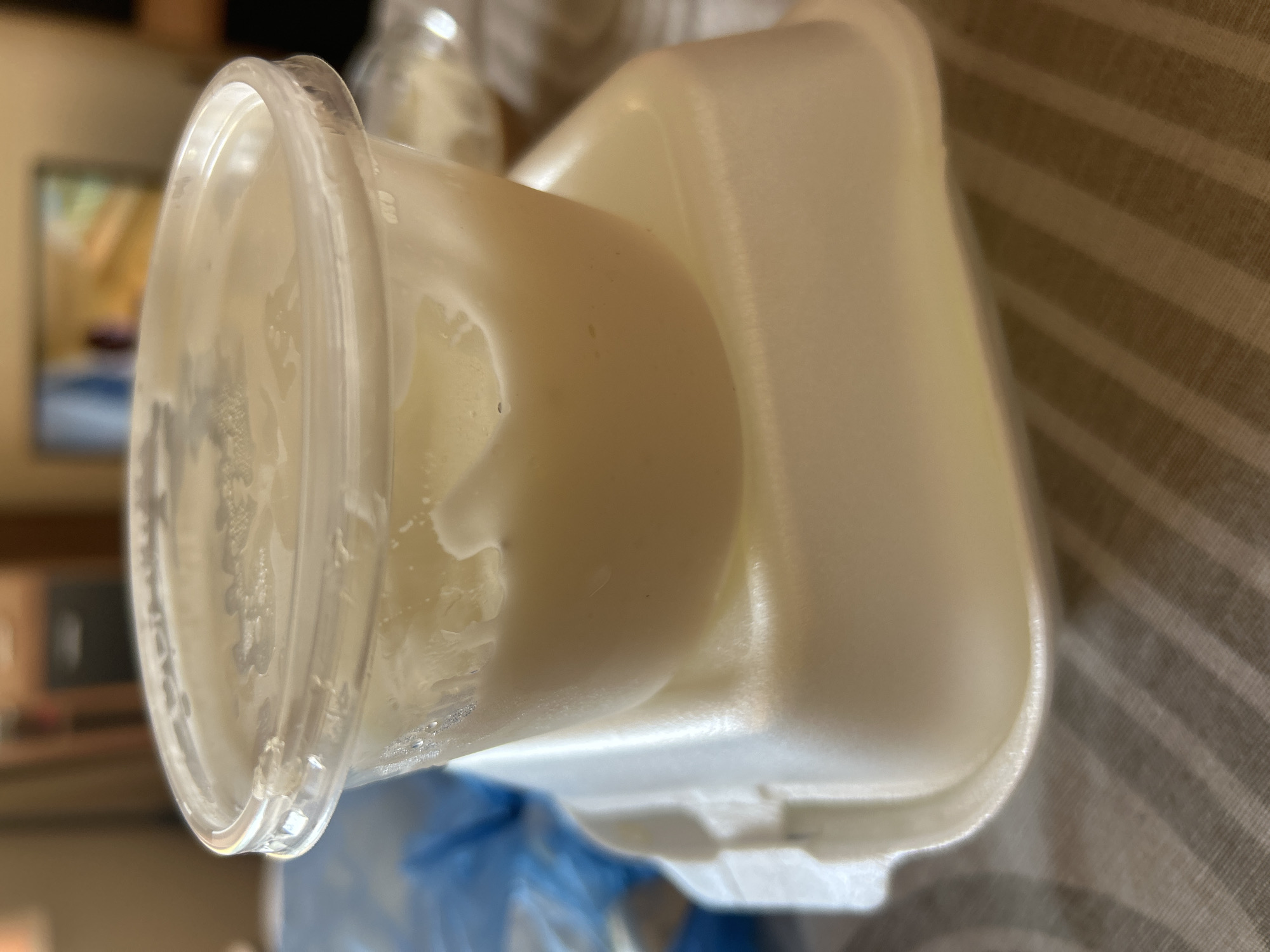 The image shows a close-up, tilted view of a white styrofoam container holding a creamy, light-colored substance, possibly food or drink. The container is partially covered by a clear plastic lid, which is also tilted. The substance inside appears to be somewhat viscous, with some lighter specks and possibly some condensation on the inside of the plastic lid.
The styrofoam container has a distinct shape with rounded edges and appears to be a disposable food container. The background is blurred, but a striped surface, possibly a tablecloth or fabric, is visible. In the further background, a hint of a television screen or a framed picture is visible, displaying muted colors.
No people are visible in the image. The lighting suggests an indoor setting, possibly during the day or evening. There is no visible text in the image. The overall impression is a still life of a food or drink container, likely in a domestic setting. The image's composition focuses on the texture and form of the contents and container.