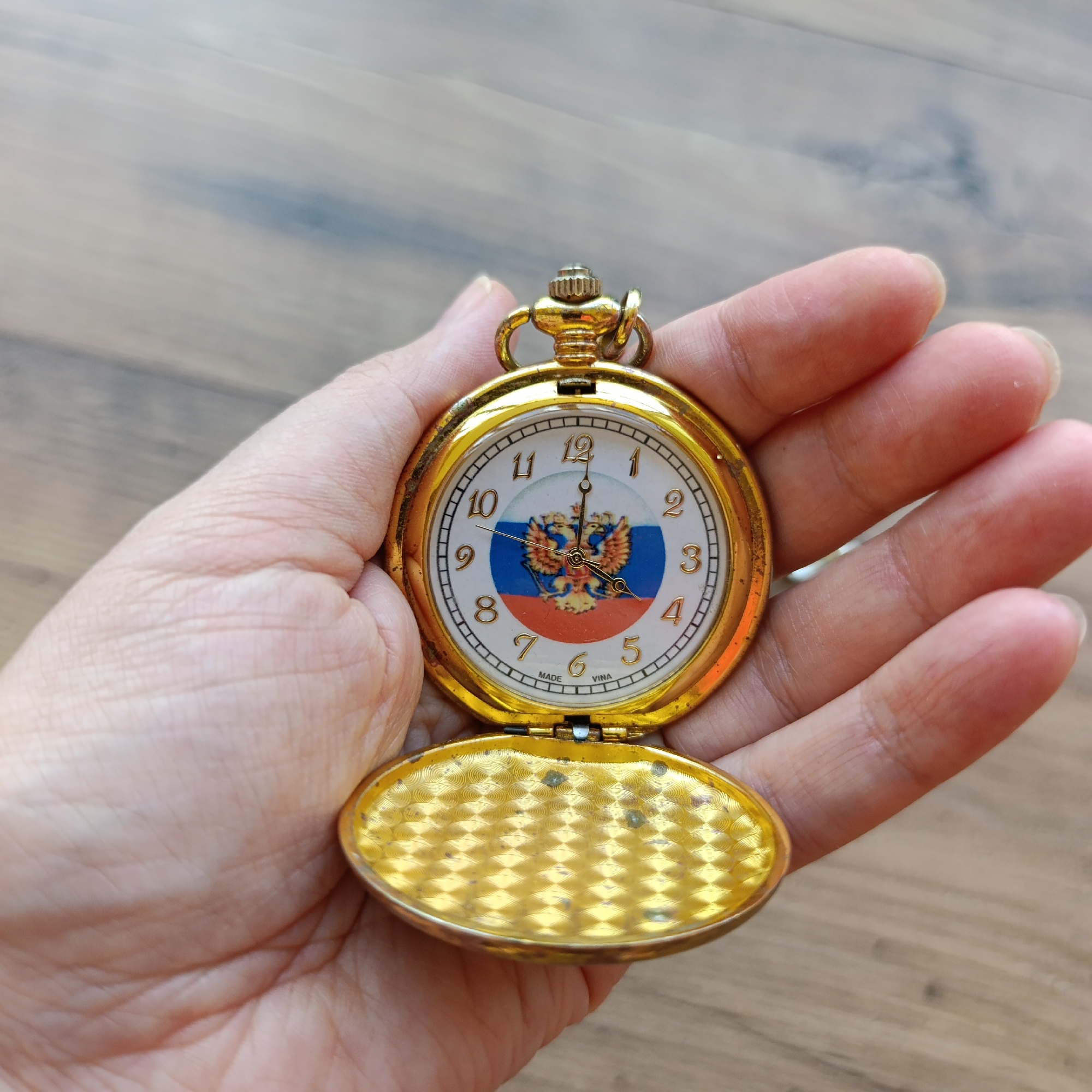 An open pocket watch with a gold-colored casing is held in a person's hand. The watch face features the Russian coat of arms with a double-headed eagle and the Russian flag colors. The watch is resting on a wooden surface. The scene appears to be indoors. The image does not provide any information about the specific location in the city of Марковский, Russia.