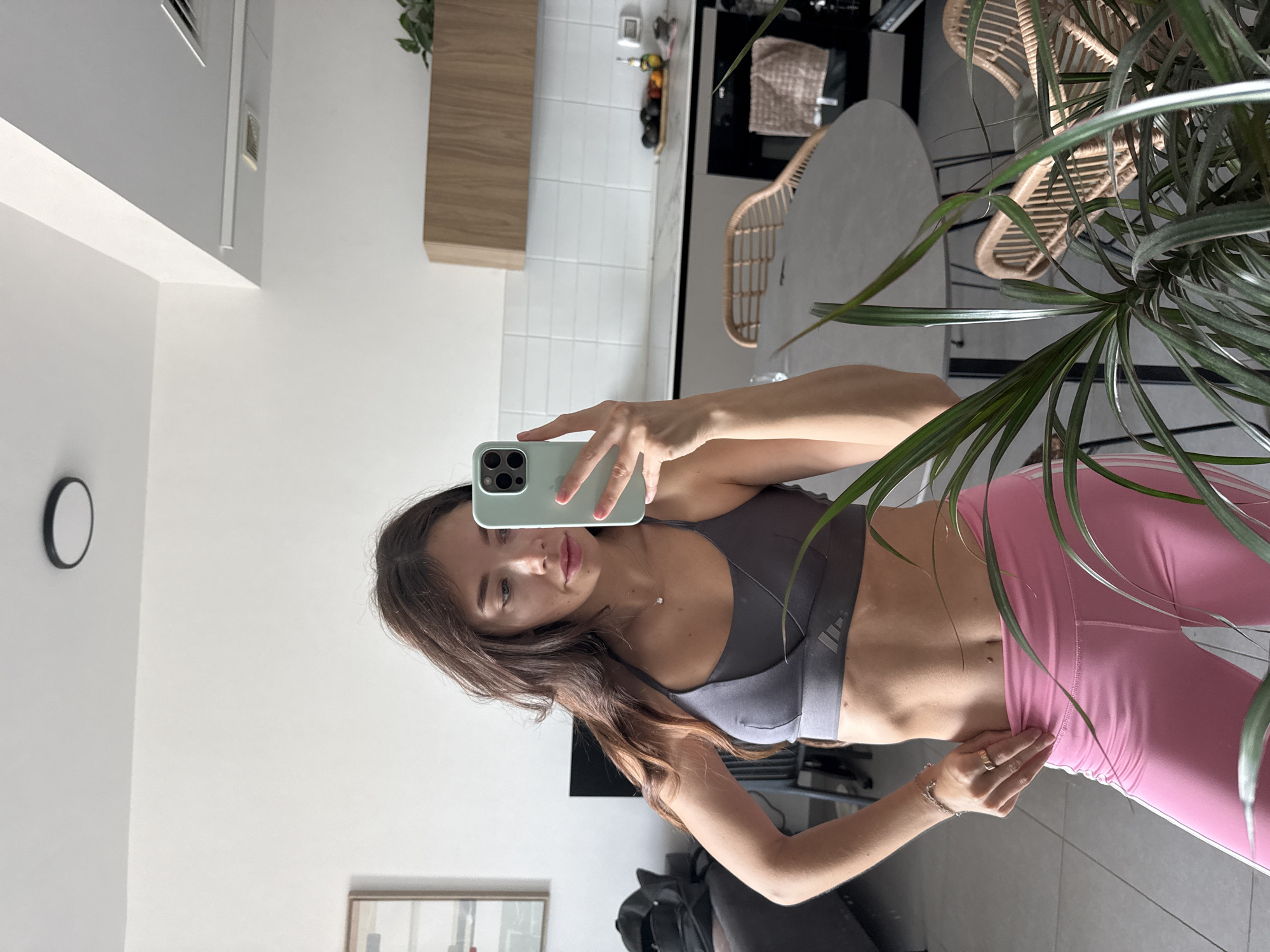 A woman is taking a selfie in a bright, modern kitchen. She is wearing a grey sports bra and pink leggings, and is holding a smartphone with a green case. The kitchen has white walls and tile backsplash, with light wood cabinets. A round table with woven chairs is visible in the background. A tall, leafy plant is positioned to the right of the frame. A clock is visible on the left wall. The scene appears to be indoors. This image does not provide specific location information within Tel-Aviv, Israel.