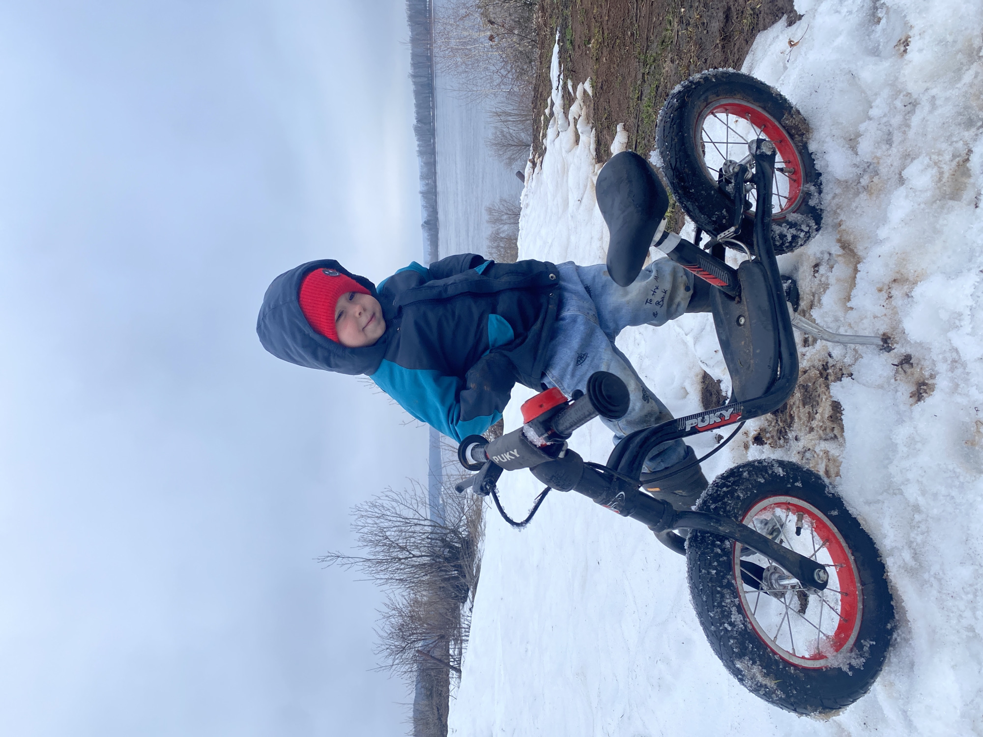 A child in a blue and black jacket and a red hat is lying sideways in the snow with a black Puky balance bike. The location is suspected to be in Ольховка, Russia. The background includes a body of water and some vegetation.