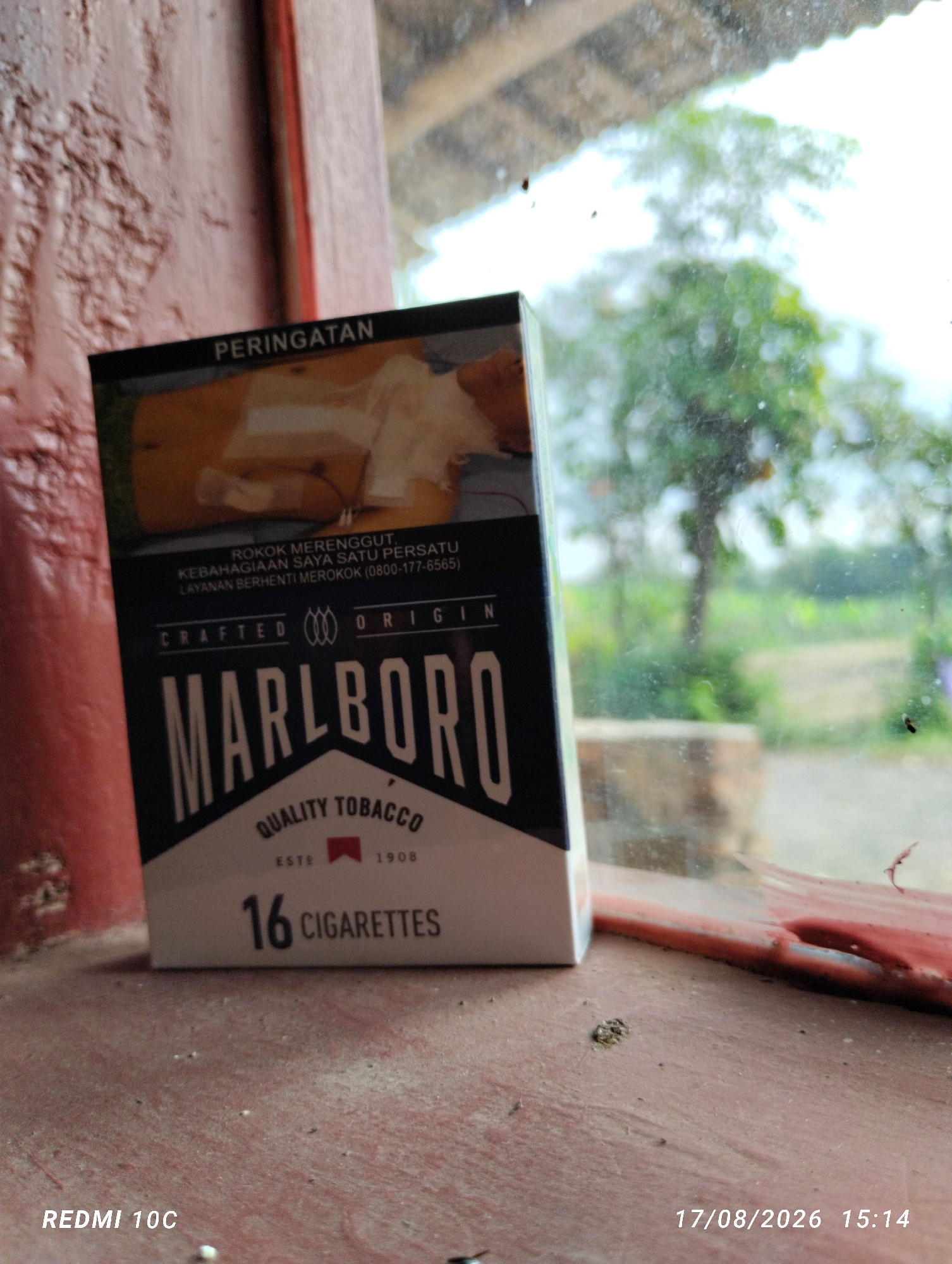 A pack of Marlboro cigarettes sits on a reddish-brown surface, possibly a windowsill, with a blurred background showing greenery and trees, suggesting an outdoor or semi-outdoor setting. The cigarette pack is the primary subject and is positioned in the lower-left quadrant of the frame. Text on the pack indicates "MARLBORO," "QUALITY TOBACCO," "ESTE 1908," and "16 CIGARETTES." There is also a warning message in Indonesian. The image includes a timestamp and device information in the lower corners: "REDMI 10C" and "17/08/2026 15:14." The overall scene appears to be captured in Kembiritan, Indonesia.