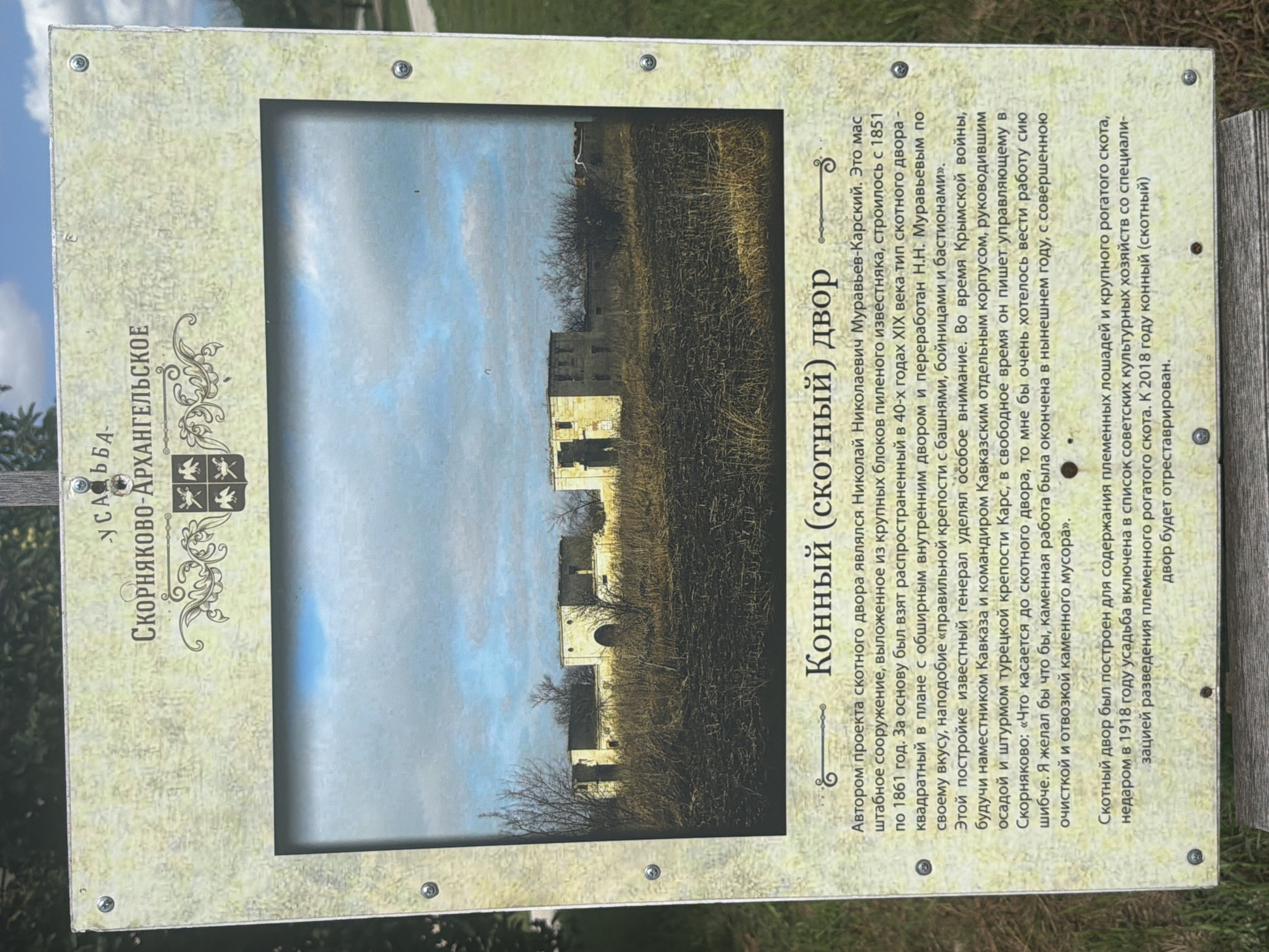The image shows an information board with a photograph of an old stone structure in a rural setting. The board has text in Russian. The photograph depicts a series of connected, dilapidated buildings with arched doorways and openings. The structures appear to be made of stone and are surrounded by dry, overgrown vegetation. The sky above is partly cloudy. The information board itself is rectangular with a textured, light-colored background and is framed with a metallic border. The text on the board is predominantly in Russian, with a title that appears to be "Конный (скотный) двор" (Horse (livestock) yard). The text below the title provides historical information about the construction and purpose of the building. The board is mounted outdoors, as evidenced by the natural lighting and surrounding greenery. The location is identified as Скорняково-Архангельское, Russia.