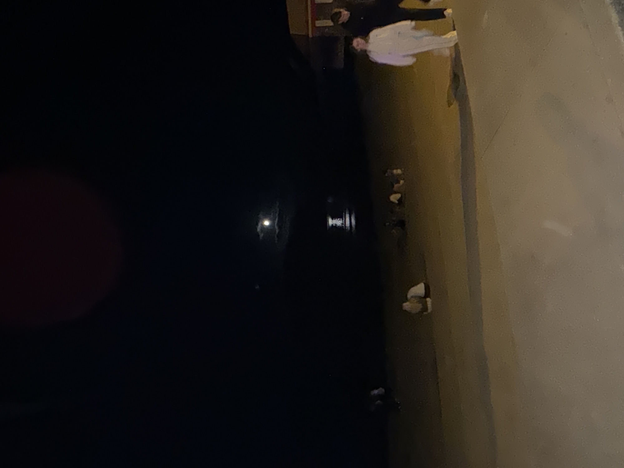 The image depicts a nighttime scene on a beach, likely the main beach in Tramore, Ireland. A full moon is visible in the dark sky, casting a faint glow on the water. In the distance, a structure or light appears on the water. On the beach, several people are scattered. In the foreground on the right, a couple walks along a paved path. The woman is wearing a light-colored hooded outfit, and the man beside her is wearing a dark jacket. Further back on the sand, a few individuals are seated or gathered, appearing to be a group of friends. The overall impression is a casual evening gathering on the beach.