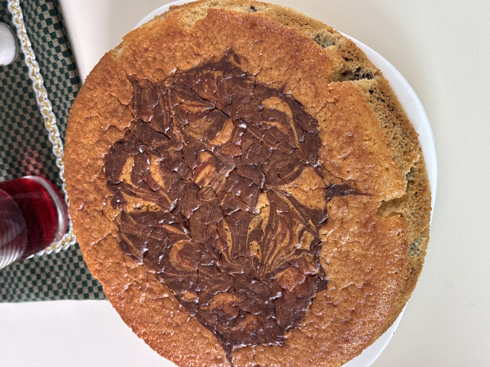 A top-down shot captures a marble chocolate chip cookie cake on a white plate. Adjacent to the cake, a glass filled with a red liquid sits beside a green and white checkered cloth with decorative trim. The scene is set against a white surface.