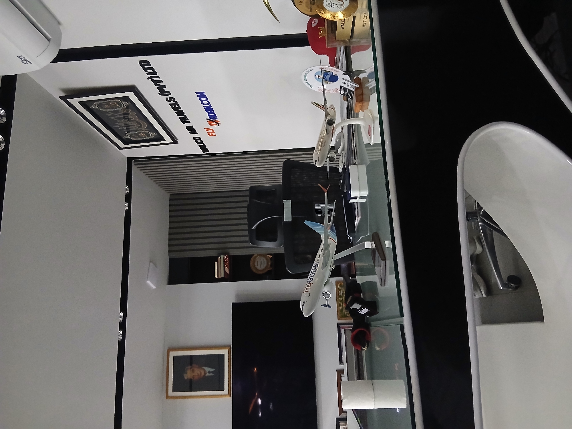An interior view of what appears to be a travel agency office in Quetta City Tehsil, Pakistan, is depicted. The scene is dominated by a modern desk with a reflective glass surface, showcasing various objects.
On the desk, there are two distinct model airplanes, suggesting a focus on aviation or travel. One model is predominantly white with blue accents on its tail, while the other is a lighter greyish-white. Alongside these models, several trophies and awards are visible, including golden-hued pieces and one with a red cap. Visible text on these awards includes "MOST IMP," "WALIC," and "MYTICO," possibly denoting achievements or recognition. A small standee with "sland" and "200" is also present, along with a roll of white paper and other small items.
Behind the desk, mounted on a white wall, large dark text spells out "WLICO AR TRAVELS (PVT) LTD" and "FLY VADAN.COM," clearly identifying the business. Below this, a framed dark picture with some text is visible. On an adjacent wall, a large black screen or television is mounted, and below it, a framed portrait of a person is displayed. A black office chair is positioned at the desk, and striped blinds cover a window or opening in the background.
Reflections of the ceiling and an air conditioner unit (with "Star" visible on it) are noticeable on the glass desktop and a dark reflective surface to the right. The overall setting is professional and organized, characteristic of a business environment.