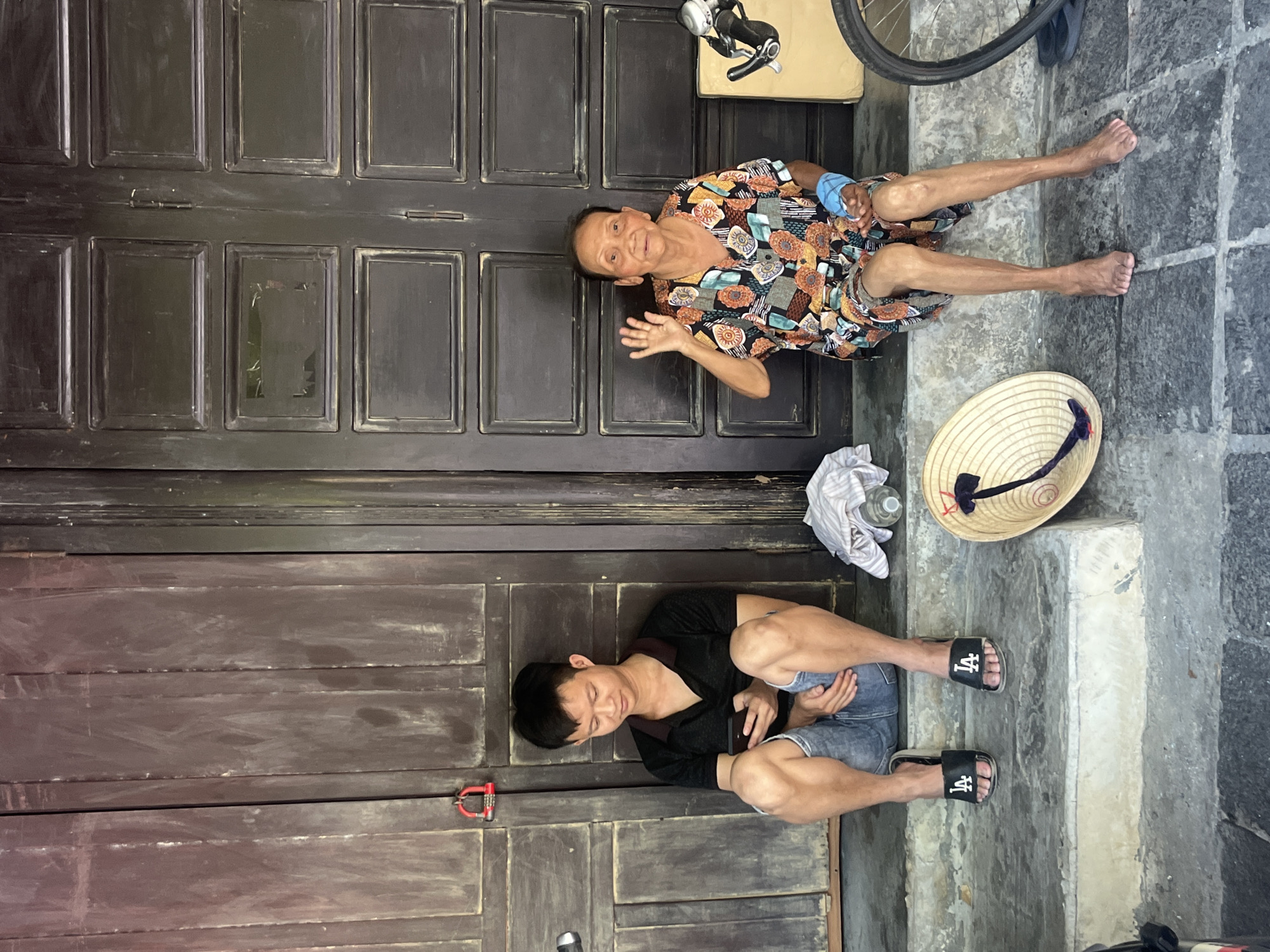 A vibrant street scene in Hội An Ward, Vietnam, depicts two individuals resting against a dark, paneled wooden building facade under bright daylight.
In the upper section of the image, an elderly woman sits on the ground, leaning against the wooden door. She is smiling warmly and waving towards the camera with her right hand. She wears a colorful, patterned short-sleeved shirt and light-colored shorts, with bare feet. A light blue bandage or wrap is visible on her left wrist.
Below her, a younger man is seated on light grey concrete steps, also against the wooden background. He is wearing a black t-shirt, denim shorts, and black slides with the white text "LA" clearly visible on the straps. He is engrossed in a black smartphone he holds in his hands, looking down at the screen.
On the steps near the elderly woman's feet, a traditional Vietnamese conical hat (nón lá) with a black and red chin strap is placed. A clear plastic water bottle and a crumpled white cloth are also on the steps. In the background, partially visible, are components of a bicycle, including a wheel and handlebars, suggesting a parked bike. A small red object, possibly a bicycle pedal or lock, is near the young man's feet.
The bright, natural lighting indicates a sunny day. The scene captures a moment of casual rest, with the elderly woman engaging friendly with the camera while the younger man is absorbed in his phone, characteristic of everyday life in the region.