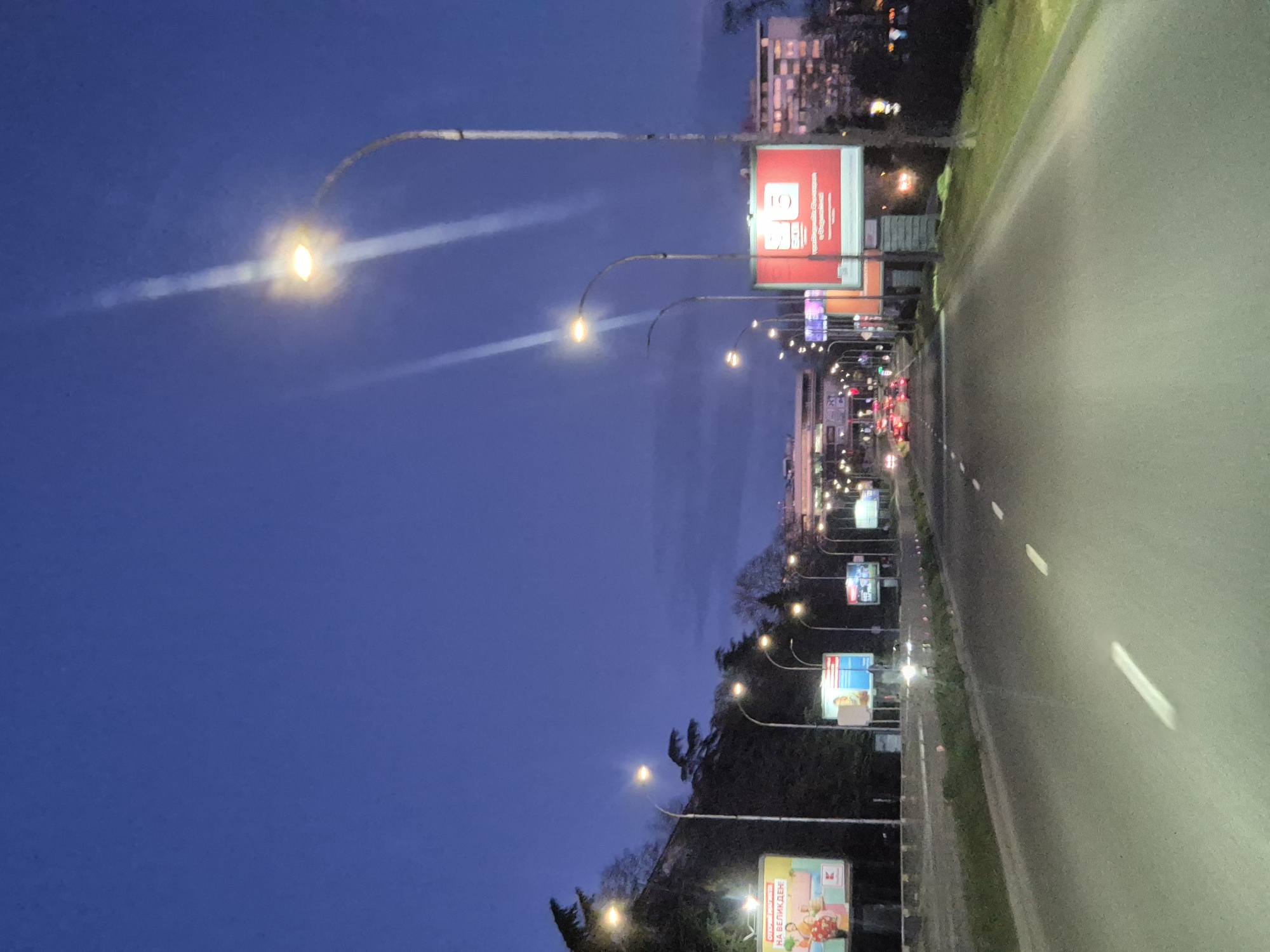 The image depicts a street scene in Varna, Bulgaria at dusk or night. A multi-lane road dominates the right side of the image, with white lane markings visible. Along the left side of the road are a series of streetlights with curved arms, illuminating billboards. The billboards display a range of advertisements, some with red, white, and blue colors. In the background, there are trees and buildings, suggesting an urban or suburban environment. The sky is a deep blue. There appear to be some cars and other light sources further down the road.