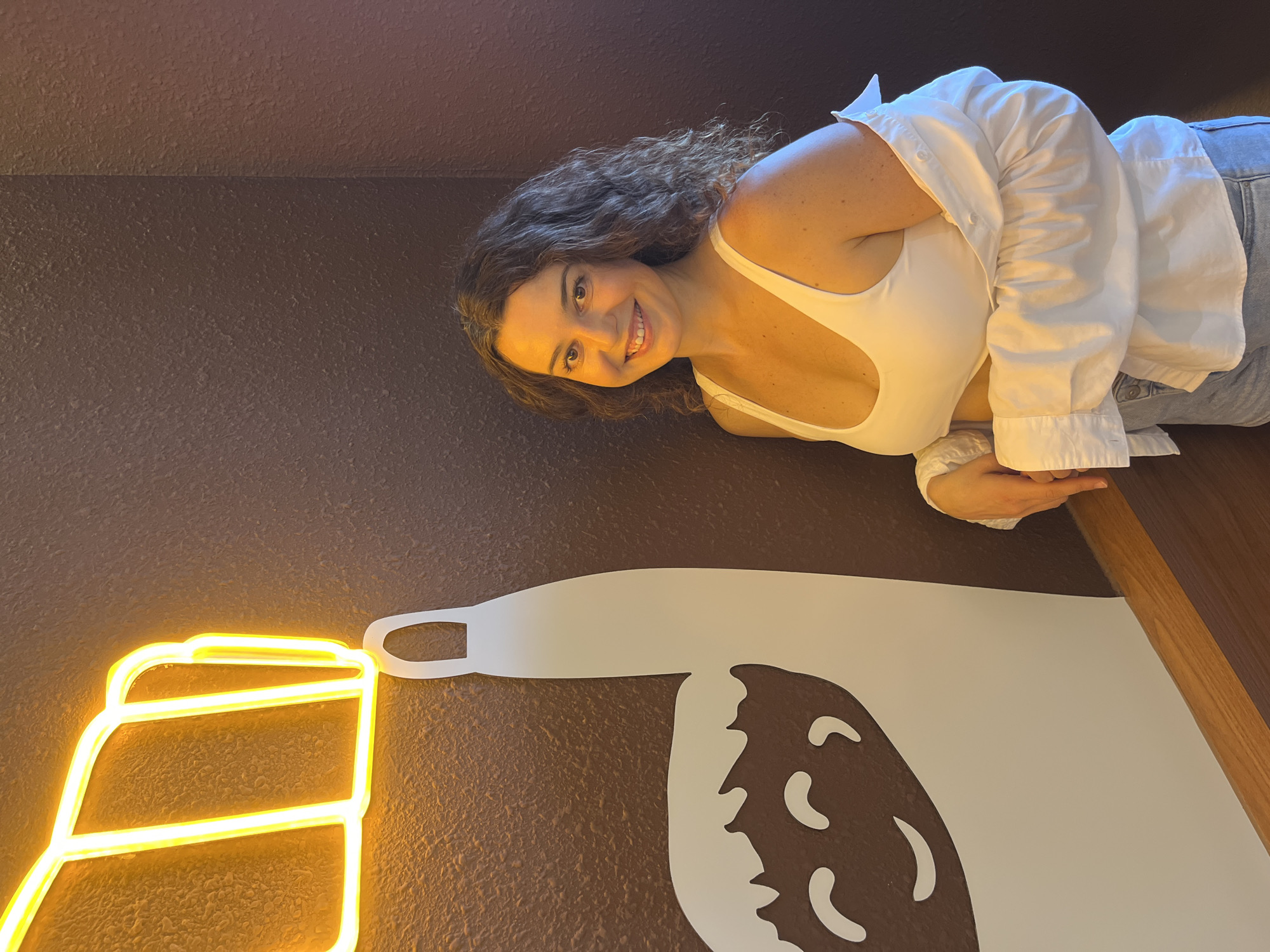 A young woman with curly brown hair and a bright smile stands in front of a dark brown wall. She is wearing a white tank top, a white unbuttoned shirt, and blue denim shorts. A neon yellow light fixture shaped like a beer mug is visible on the left side of the image, partially obscuring a white cartoon llama illustration. The llama has a simple, smiling face. The background wall has a textured appearance. The woman is leaning slightly against the wall and the llama illustration. The overall scene suggests an indoor setting, possibly a restaurant or bar, in Phu Thuy Ward, Vietnam.