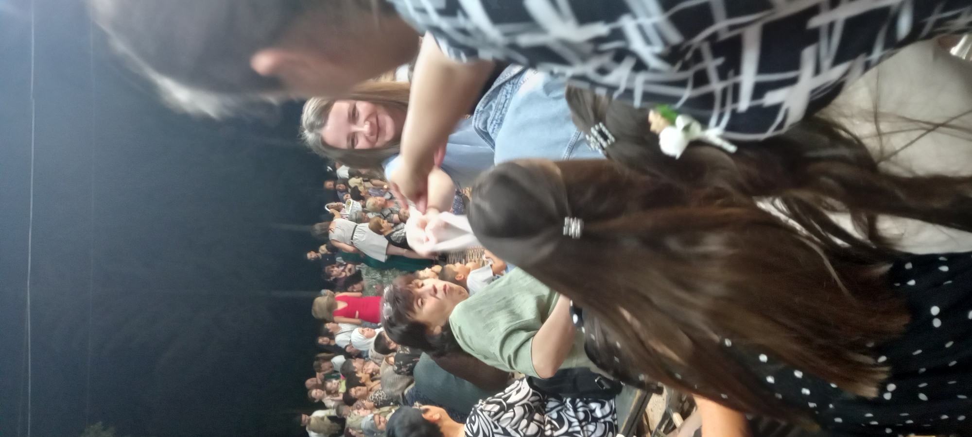 The image shows a crowd of people gathered outdoors at dusk, likely for an event or performance. In the foreground, a woman with long brown hair, adorned with a decorative hair clip, is seated. To her right, another woman with shoulder-length brown hair and a smiling expression is visible, wearing a light blue top and jeans. She is interacting with someone to her right, whose arm and patterned shirt are partially in frame. Behind them, a larger group of people are seated or standing, some wearing traditional clothing. The background is dark, with trees faintly visible, suggesting a natural or park-like setting. The image is taken from a low angle, looking up at the people. The specific location in the city of Lyuben, Bulgaria, cannot be determined from the visual information provided.