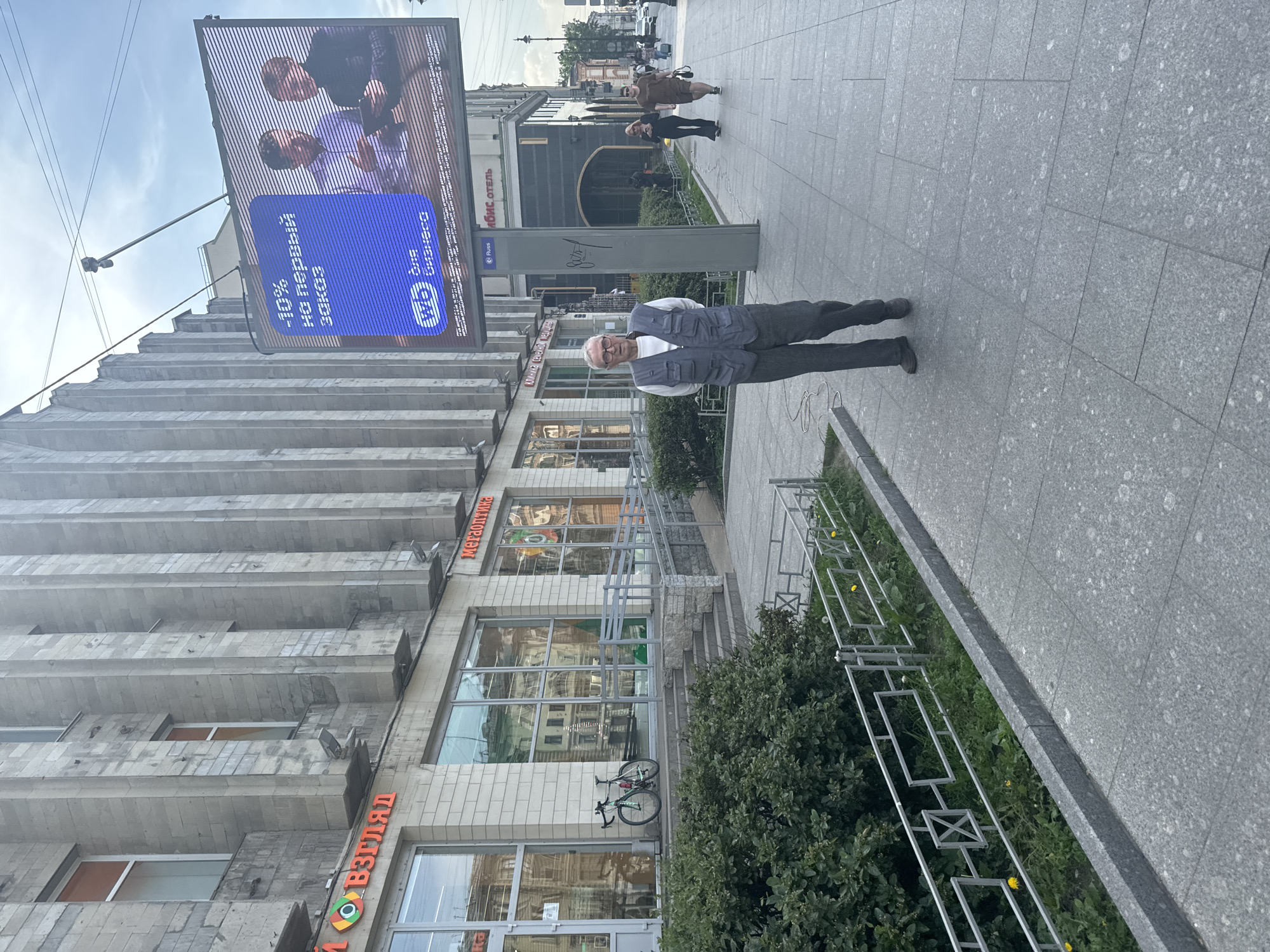 A man, appearing to be in his late 60s or early 70s, stands on a sidewalk in front of a building in Saint Petersburg, Russia. He is wearing a light-colored vest over a white shirt and dark trousers, with glasses on. The building features a light-colored stone facade with vertical architectural elements and multiple windows. A large digital billboard is mounted on the building, displaying an advertisement with Russian text and an image of two men. The sidewalk is paved with grey stone tiles, and there is a metal railing and greenery alongside it. In the background, other pedestrians are visible, and a street with cars can be seen. The sky is overcast. The signs on the building appear to be for shops or businesses. The overall scene depicts a typical urban streetscape in Saint Petersburg.
