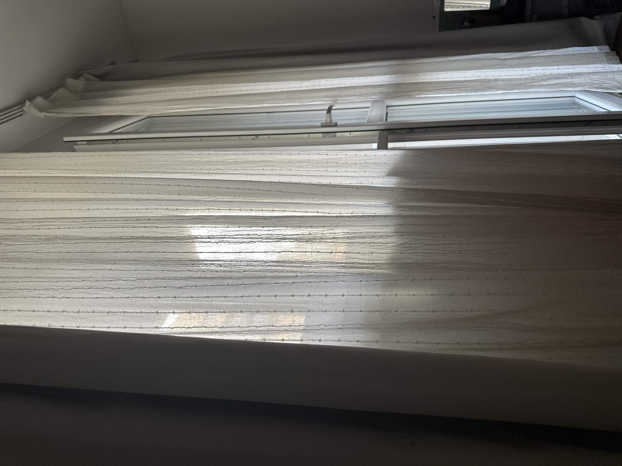 The image shows a close-up view of sheer white curtains with subtle vertical patterns and small raised dots. The curtains are partially drawn, revealing a partially open white-framed window with a silver handle. Sunlight filters through the sheer fabric, casting a soft glow and creating shadows. On the left, a darker, opaque curtain is visible, suggesting a layered window treatment. The setting appears to be indoors, with a hint of a wall and ceiling visible at the top. The overall impression is one of a domestic interior, likely a room with a window. The context of Sofia, Bulgaria, is not discernible from the visual information provided in the image.