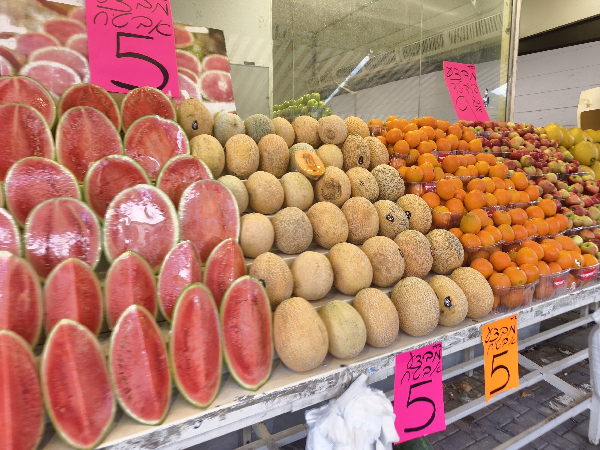 The image shows a fruit stand in Petah Tikva, Israel. The stand is filled with various types of fruit, including sliced watermelons, cantaloupes, oranges, apples, and pears. The fruits are arranged in rows and some are sold in clear plastic containers. Pink and orange signs are visible with the price '5' written in Hebrew script. The stand itself appears to be made of metal and is situated outdoors.