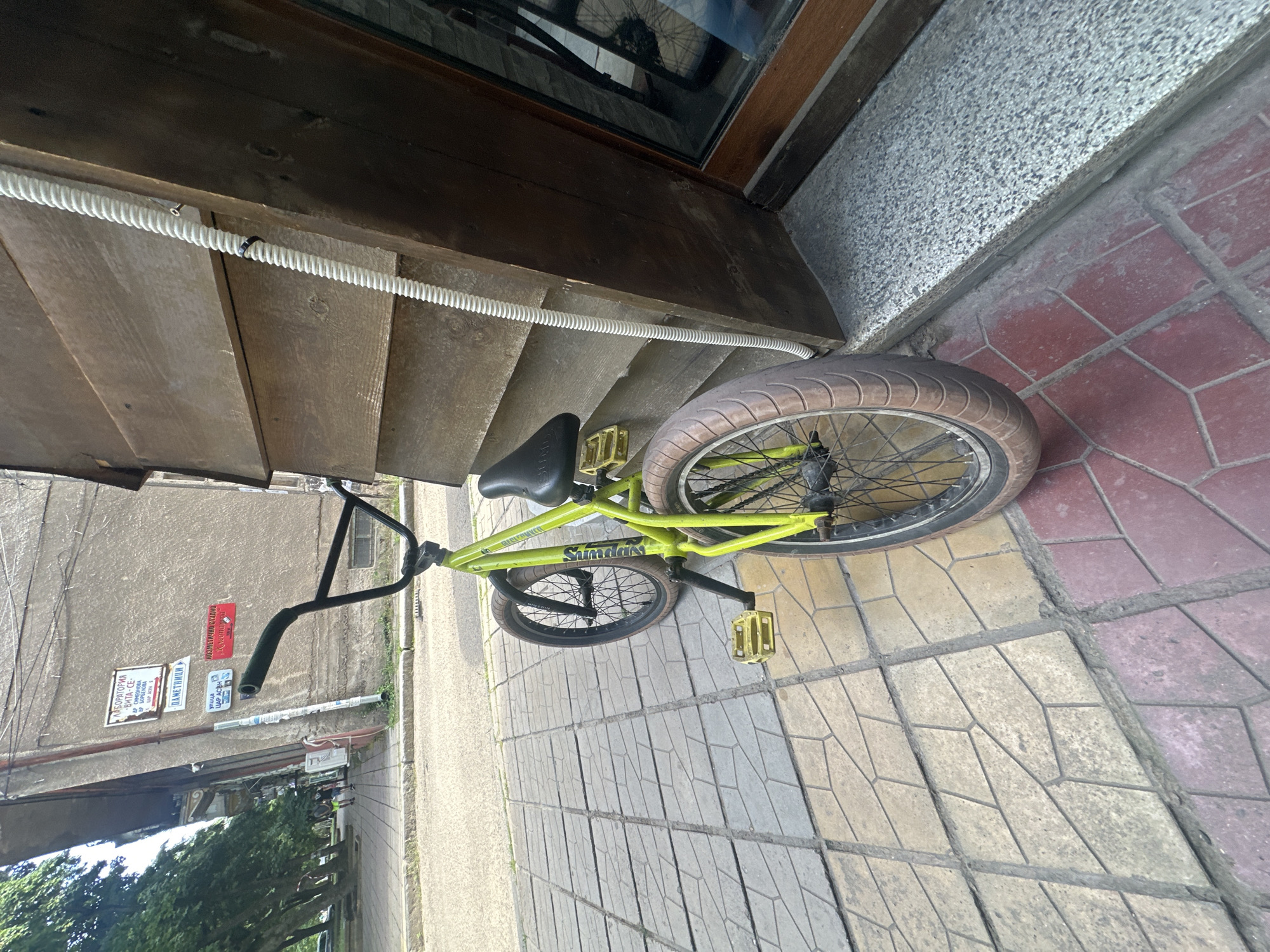This image depicts an urban street scene located on Tsar Asen Street in Lom, Bulgaria, as indicated by Cyrillic signage visible on a building. The primary subject is a bright lime green BMX-style bicycle, branded "Sunday" on its downtube and "Odyssey" on its front fork. The bicycle, equipped with black handlebars, a black seat, and yellow square pedals, is resting vertically against a dark brown, weathered wooden facade composed of horizontal planks. Its wheels feature black rims, black spokes, and tires with brownish-tan sidewalls and black treads; the rear tire is notably wider.
To the left of the bicycle stands a multi-story building with a light grey, textured exterior, characterized by a prominent slanted architectural section on its left side. Several Cyrillic signs are affixed to this building: "ЛАБОРАТОРИЯ -ВИТА-СЕ-" (Laboratory -Vita-Se-), "ДР СИМЕОНОВА БИРКОВА" (Dr. Simeonova Birkova), "ПАМЕТНИЦИ" (Monuments), "УЛИЦА ЦАР АСЕН" (Street Tsar Asen), and "КОЗМЕТИЧНО СТУДИО ЕЛЕГАНС" (Cosmetic Studio Elegance).
The ground directly in front of the wooden facade is paved with red hexagonal/geometric tiles, transitioning into yellowish-grey irregularly shaped paving stones that extend towards the left. Beyond the tiled sidewalk, a grey concrete curb separates it from an asphalt road. A white corrugated conduit or cable runs vertically along the right side of the wooden facade, secured by a black clamp. To the far right of the wooden structure, a dark brown-framed window is visible, reflecting external light. In the distant background on the left sidewalk, two to three indistinct human figures are walking away from the camera. Green deciduous trees line the left side of the street. The scene is illuminated by natural daylight.