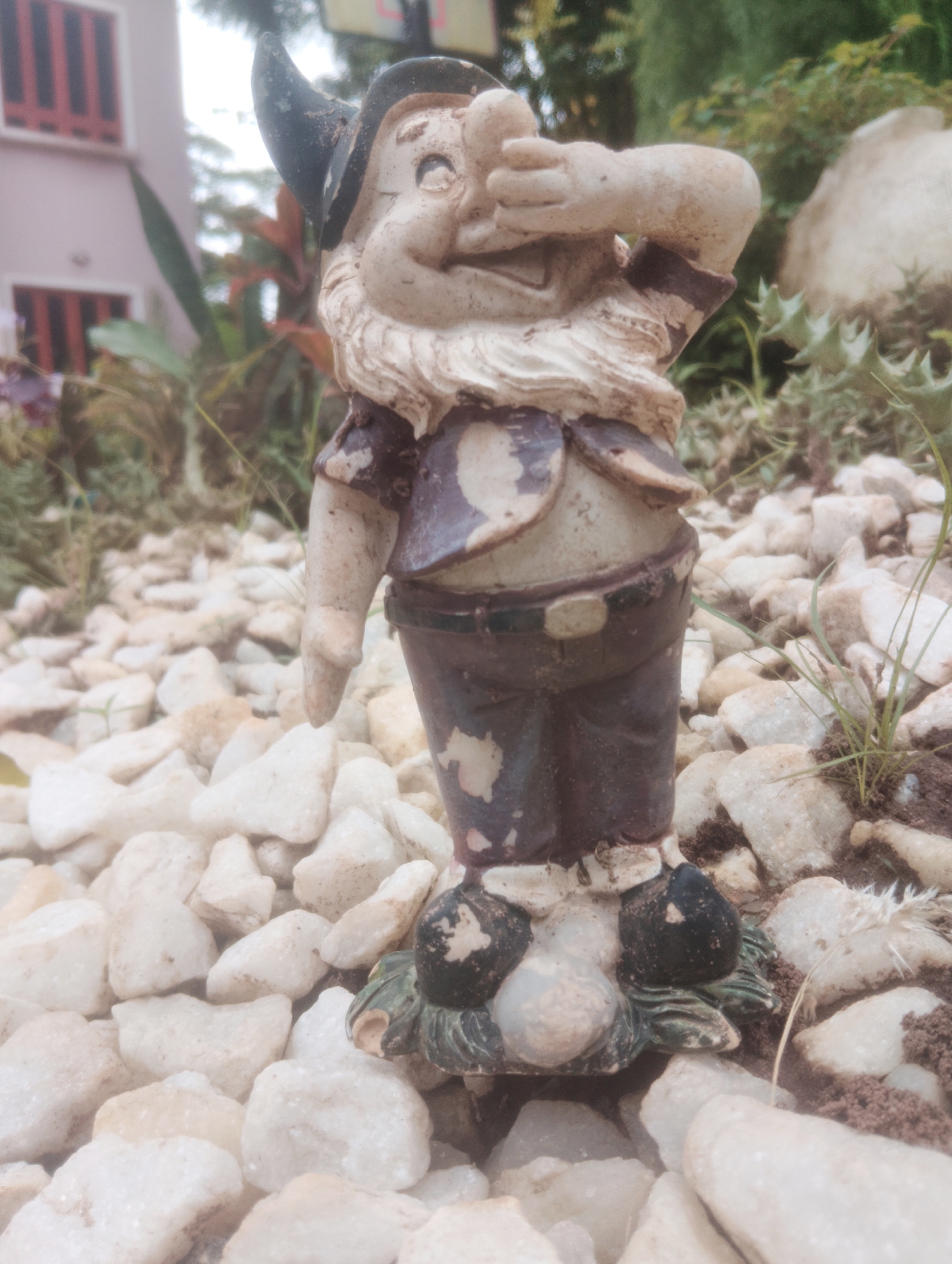 In Aketu-Oja, Nigeria, a weathered garden gnome stands amidst white gravel. The gnome, with a pointed hat and a beard, holds its hands to its face, suggesting a gesture of distress or surprise. Its clothing, painted in shades of brown and blue, shows signs of wear and tear. The background is softly blurred, revealing glimpses of a pink building with red-barred windows and lush green foliage, indicative of a domestic garden setting. The overall scene is serene, with the gnome as the central focus against the natural backdrop of a Nigerian garden.