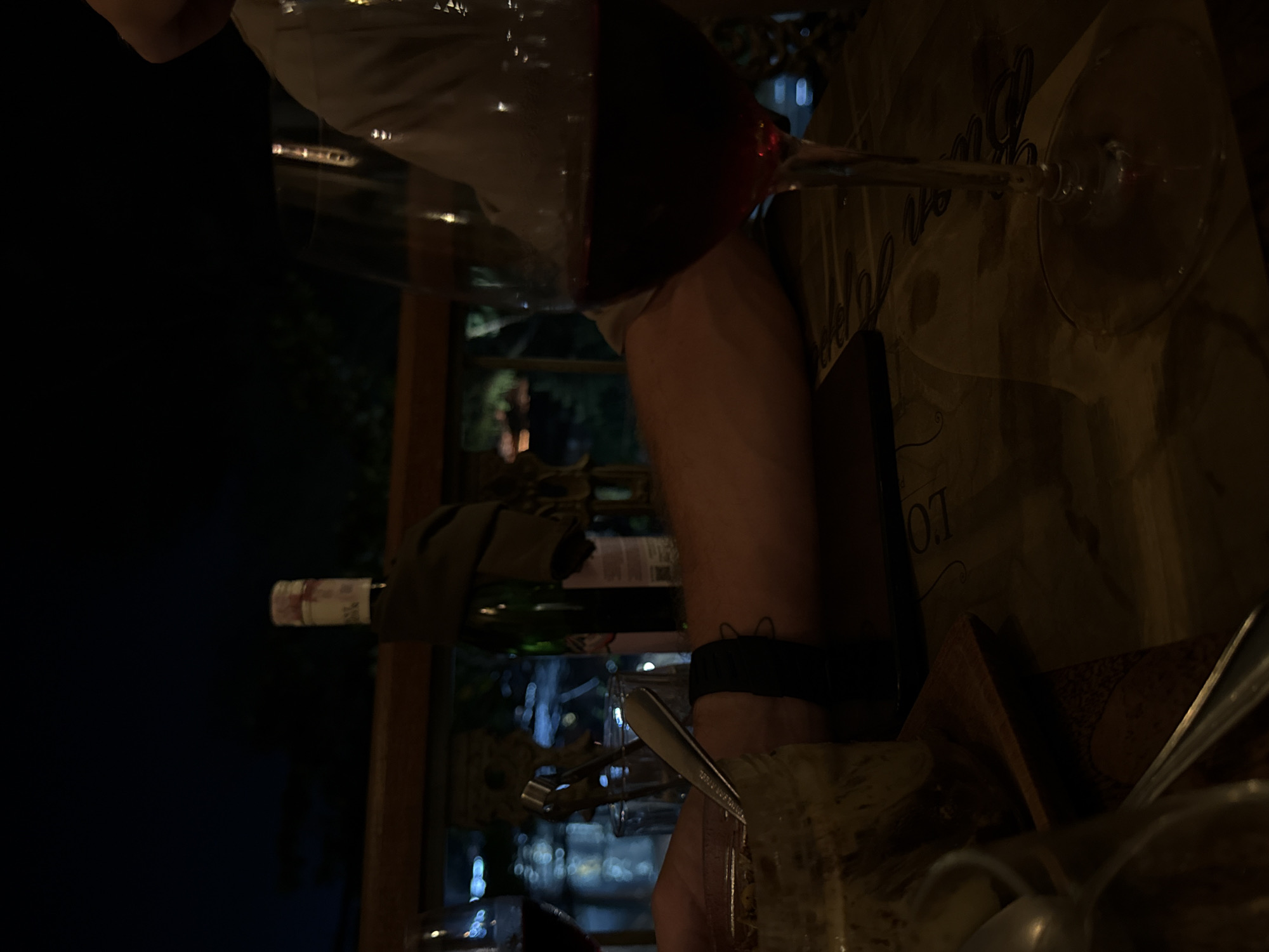 The image, oriented vertically in its original form, depicts a dimly lit indoor scene, likely a restaurant or bar, at night. The central focus is a person's arm, wearing a dark wristband, holding a wine glass filled with red wine, tilted as if being poured or moved. The liquid inside the glass reflects the surrounding low light.
In the foreground, another empty wine glass, with its stem and wide base, rests on a table covered with a textured, light-colored material, possibly a paper placemat or tablecloth. The letters "LO" are distinctly visible on this surface, suggesting part of a word like "LONDON" or a brand name. To the right, a collection of utensils, including a spoon and a pair of tongs, are visible, partially obscured.
A green wine bottle with an indistinct label is positioned in the mid-ground, suggesting it's part of the dining setup. Nearby, a folded napkin or cloth drapes over an unseen object. The background is dark, likely indicating nighttime outdoors through a window or a very dimly lit section of the interior. Soft, out-of-focus lights in the distance suggest an urban or populated setting. The overall ambiance is warm, intimate, and relaxed, consistent with an evening meal or social gathering involving wine.