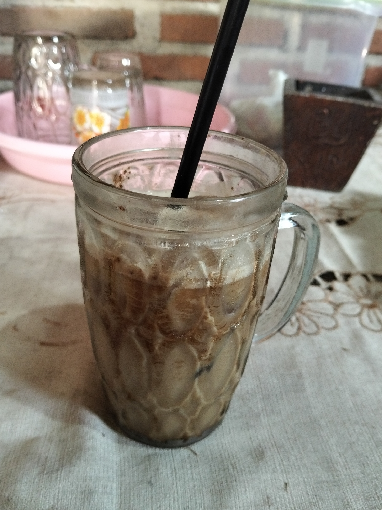 A glass of iced coffee sits on a patterned tablecloth. The glass is partially filled with a creamy brown liquid and ice cubes. A black straw extends from the top of the glass. In the background, several other glasses are visible, as well as a brick wall and a dark, wooden object. The scene appears to be indoors, possibly in a residential setting or a small cafe in Panimbang, Indonesia.
