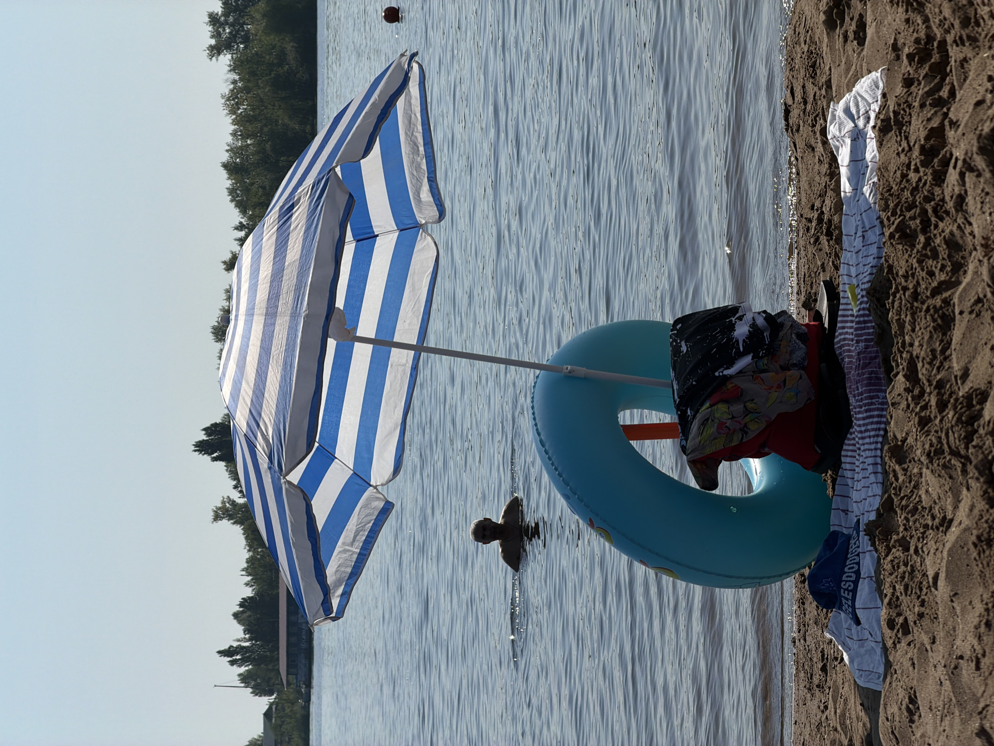A beach umbrella with blue and white stripes shades a blue inner tube on a sandy beach next to the Volga River, Saratov, Russia. A person is swimming in the river behind the inner tube. Clothes are piled on the sand near the inner tube. A blue baseball cap with the word "DODGERS" printed on it lies on a towel. Trees and buildings are visible in the distance on the opposite bank of the river. The sky is clear and blue.