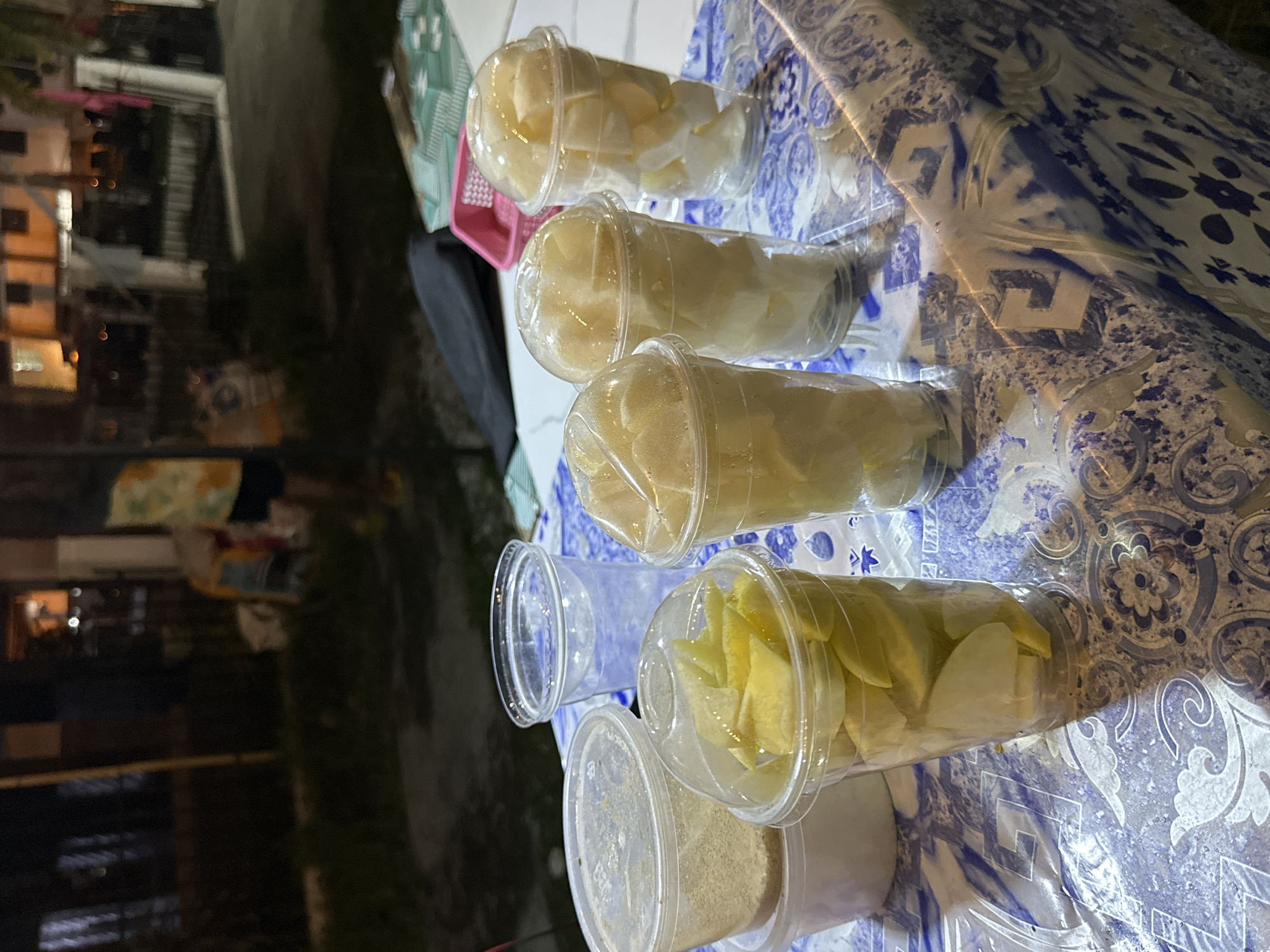 The image captures a nighttime street food scene in Davao City, Philippines. A table in the foreground is covered with a blue and white patterned plastic tablecloth. On the table, five clear plastic cups with dome lids contain cut fruit: one cup holds slices of yellow fruit, while four other cups contain chunks of white or pale yellow fruit. Adjacent to these, a lidded clear plastic container is filled with a light brown granular substance, and a single empty clear plastic cup stands nearby. A pink plastic basket and a dark bag are also partially visible on the table's edge. The background is dark and blurred, revealing faint outlines of buildings with scattered artificial lights and a partially visible person standing to the left. The ground in the background appears to be a mix of dark pavement and some sparse greenery.