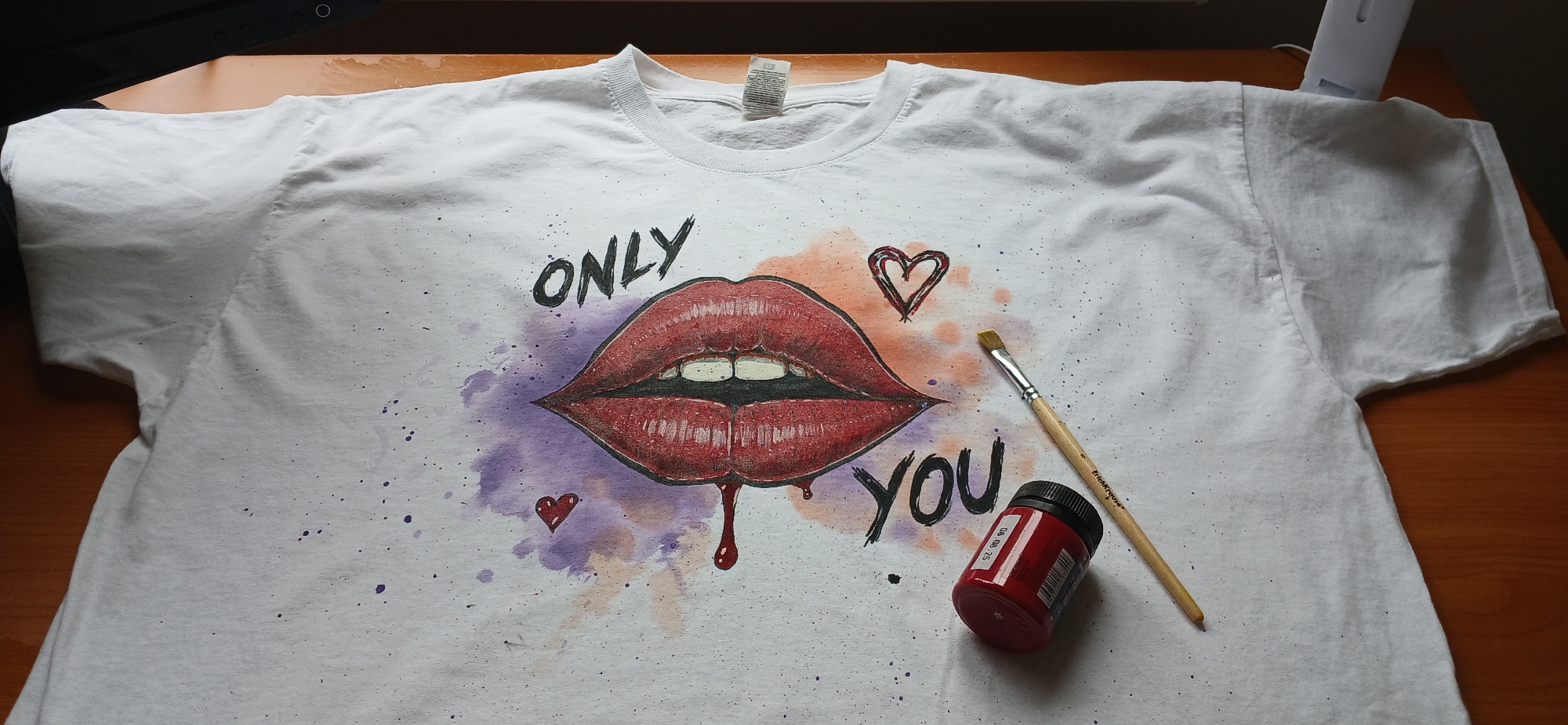 A white t-shirt with a drawing of red lips with black outlines and white teeth is centered on a brown wooden surface. Above the lips, "ONLY" is written in black. Below the lips, the word "YOU" is written in black. There are two heart drawings, one above the right side of the lips and one to the left and slightly below the lips. There is a watercolor effect of purple to the left of the lips and orange to the right of the lips. Splatters of purple and black dots are across the t-shirt. A brown-handled paintbrush and a small bottle of red paint are to the right of the lips. The t-shirt has a white tag at the neck. A black object is partially visible in the upper left corner of the image and a white object is partially visible in the upper right corner of the image. There is no indication of this image being taken in Riga, Latvia.