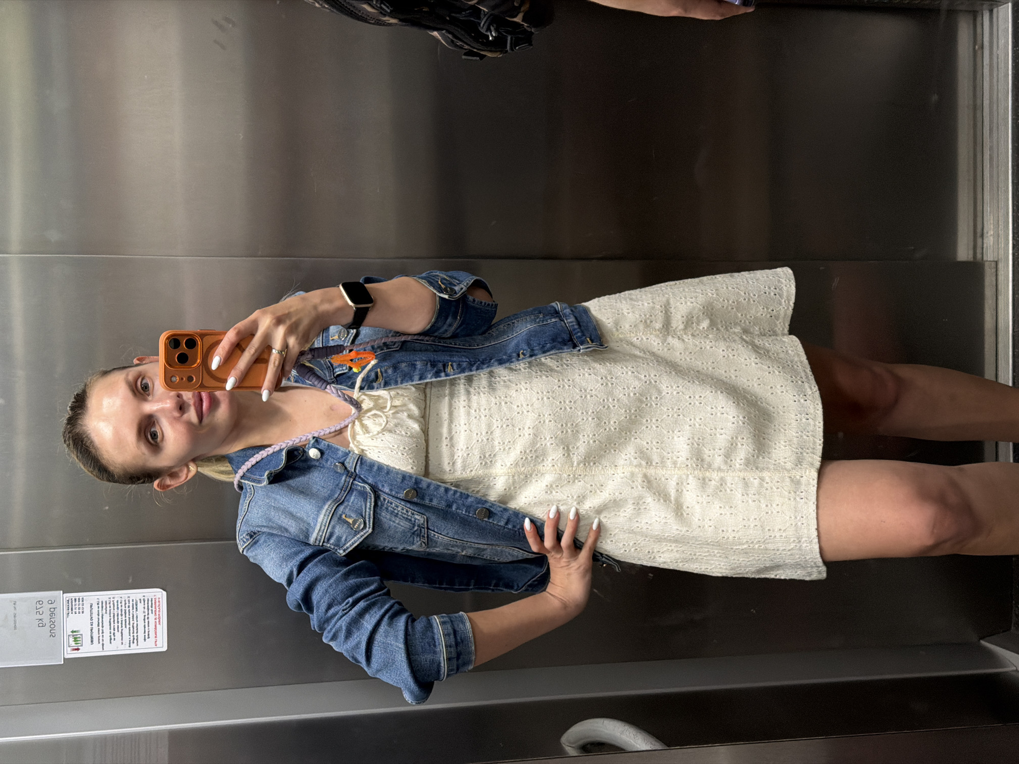 A woman is taking a selfie in what appears to be an elevator, based on the metallic walls and a safety information sticker visible in the lower left corner of the image. She's wearing a white dress and a blue denim jacket. She's holding an orange phone in her left hand to take the photo, and her right hand is resting on her hip. She has a wristwatch on her left wrist. It is not possible to identify a location in Budva, Montenegro from this image.