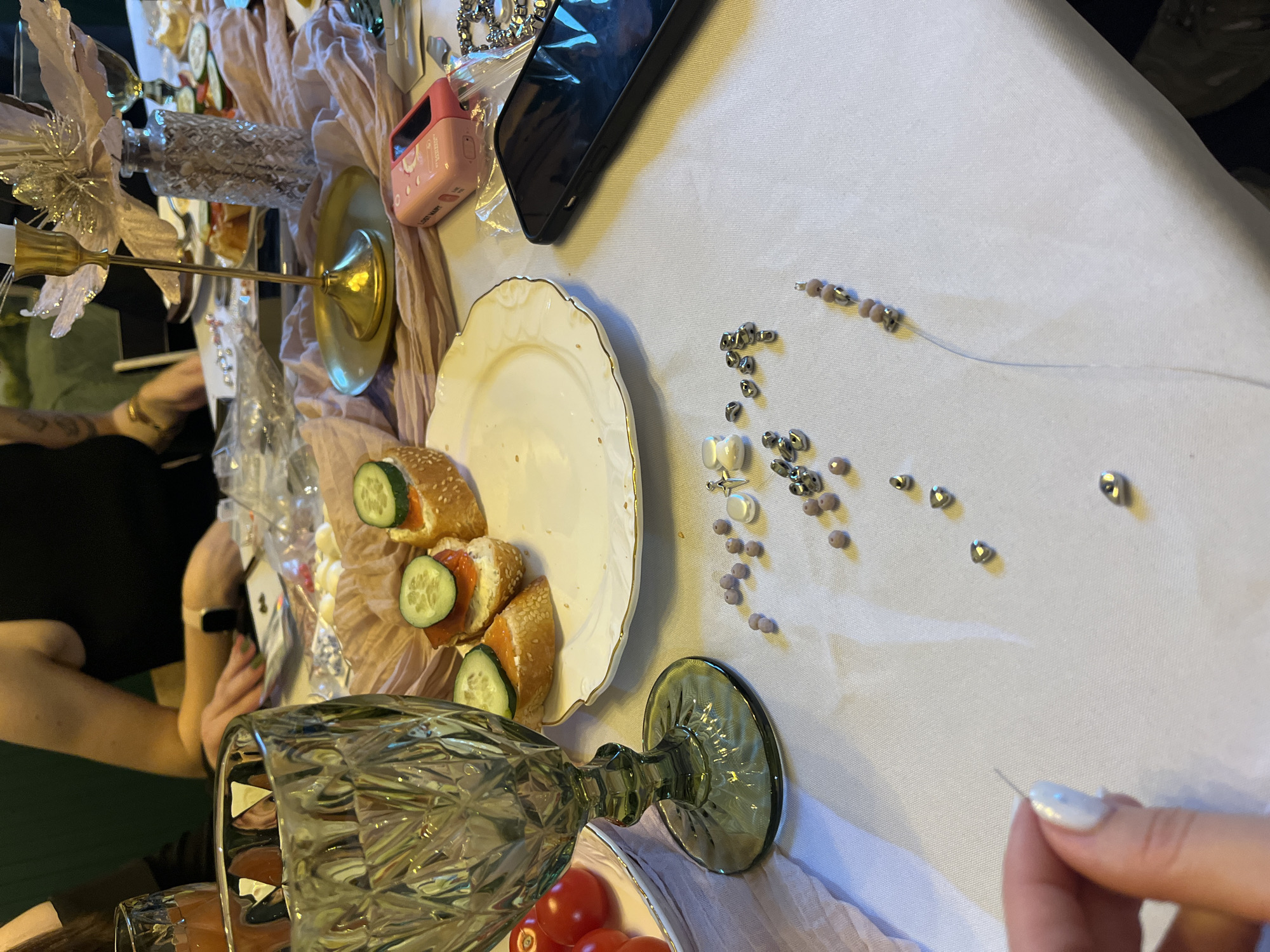 A tabletop scene is depicted with various objects arranged on a white linen tablecloth. A white plate with gold trim holds small canapés topped with cucumber slices, suggesting a catered gathering. Scattered on the cloth are beads of silver, white, and mauve hues, with a needle and thread implying the activity of jewelry making. A tall, textured green-tinted glass lies on its side near the lower edge of the frame. There is a cellphone, a pink-cased device, a decorative silver candle holder, and various other decorative elements are present. In the background, partial views of individuals are visible, suggesting they are participants in the event. The absence of landmarks and details typical of Saint Petersburg preclude confidently attributing the scene to this specific location. The scene is consistent with a craft-based activity coupled with a social event, likely indoors under either natural or artificial lighting.
