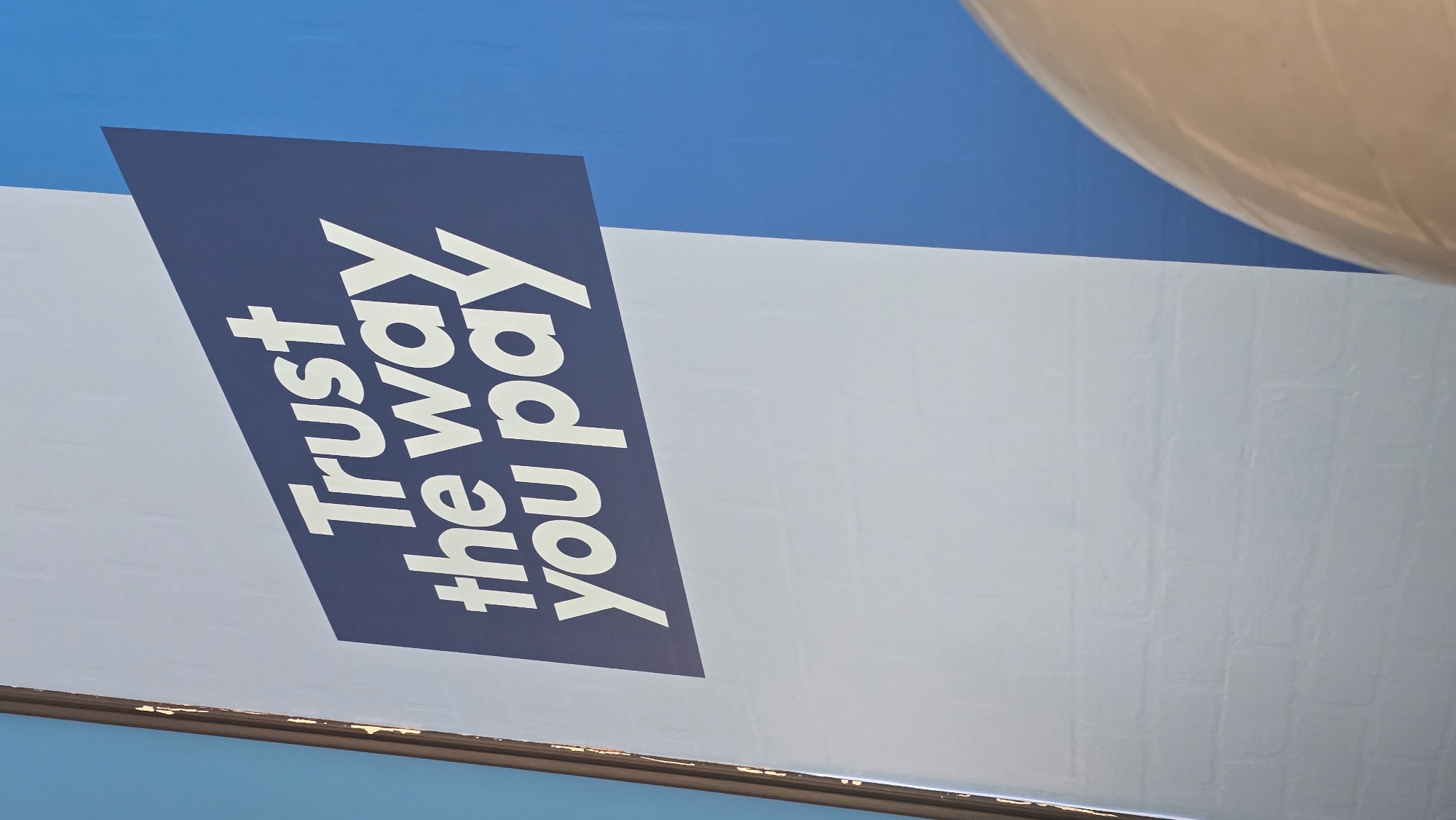 The image shows a portion of a billboard in Varna, Bulgaria. The visible part of the advertisement features the slogan "Trust the way you pay" in white text on a dark blue rectangular background. The billboard itself is mounted on a white surface. The background above the billboard is a light blue color, likely the sky. A white spherical object is visible in the upper right corner, potentially part of a building or structure. The image is taken from a low angle, looking up at the billboard.