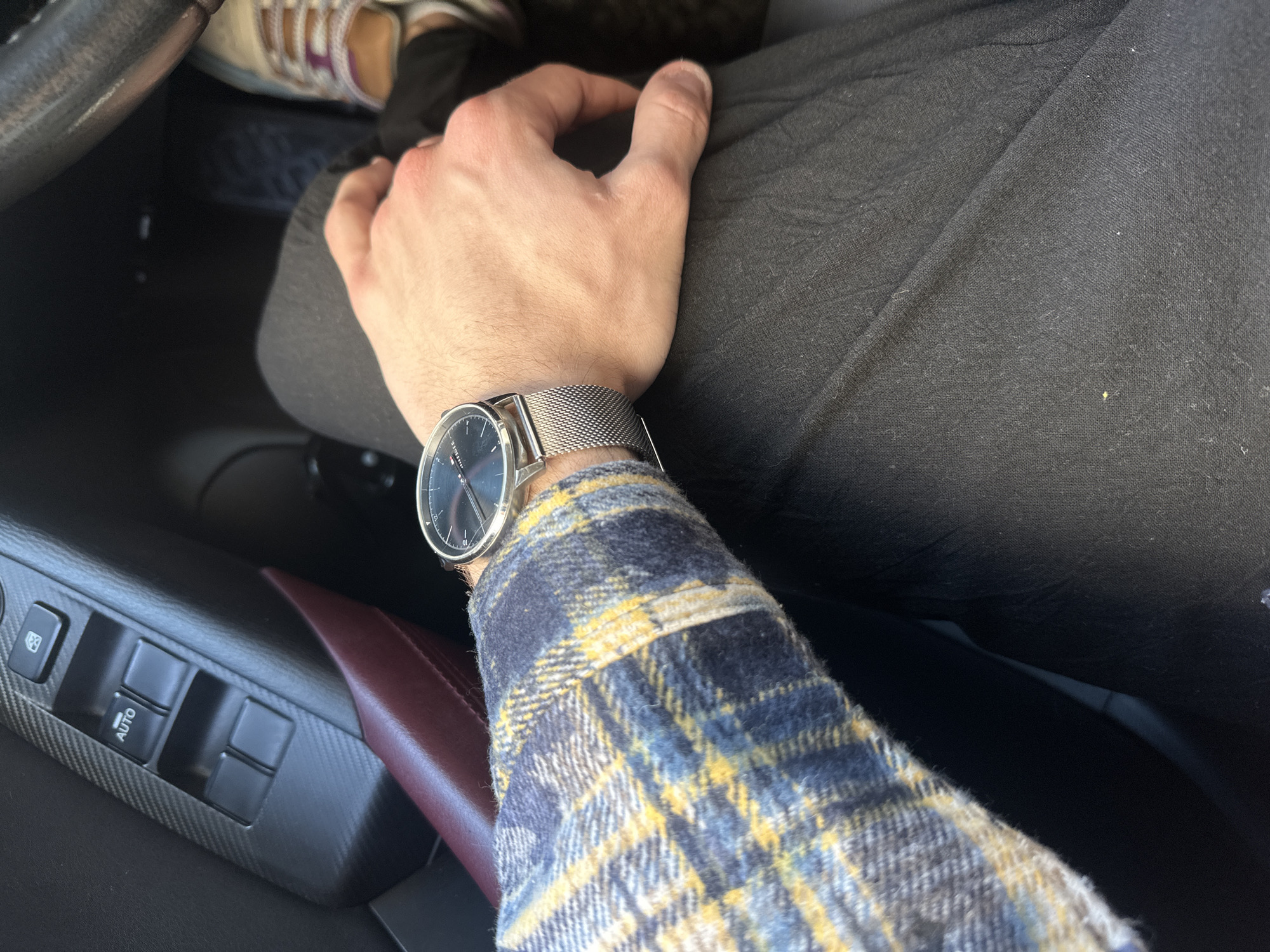 A person wearing a blue and yellow plaid shirt and a wristwatch with a silver mesh band is seated in a car. The person's hand rests on their dark grey pants. Part of a shoe, possibly a sandal, is visible in the upper part of the image, near the car's steering wheel. The car's interior features carbon fiber-patterned trim and a maroon leather detail. The controls for the car's windows are visible on the door panel. The image does not depict any location in Gabrovo, Bulgaria.