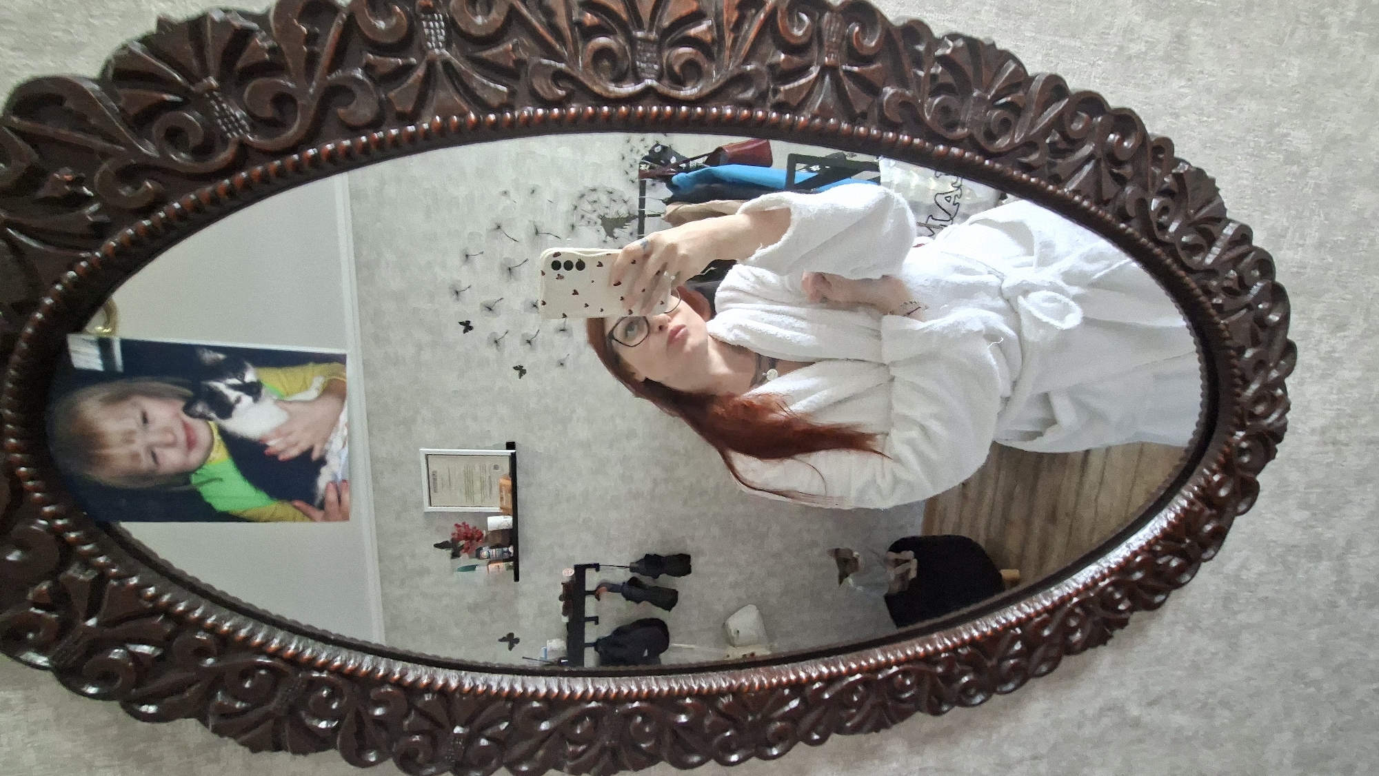 A woman with long red hair and glasses, wearing a white bathrobe, takes a mirror selfie. She is holding a smartphone in her right hand, which has a white case with a heart pattern. She is looking at the phone's reflection in a large, ornate, dark brown oval mirror. Behind her, in the mirror's reflection, a framed picture of a young child holding a black and white cat is visible on the wall. To the left, a cat is sitting on a black stool. The room appears to be a bedroom or dressing area with clothing hanging on hooks on the wall. The overall environment suggests a domestic setting. The image was taken in Myadzyel, Belarus.