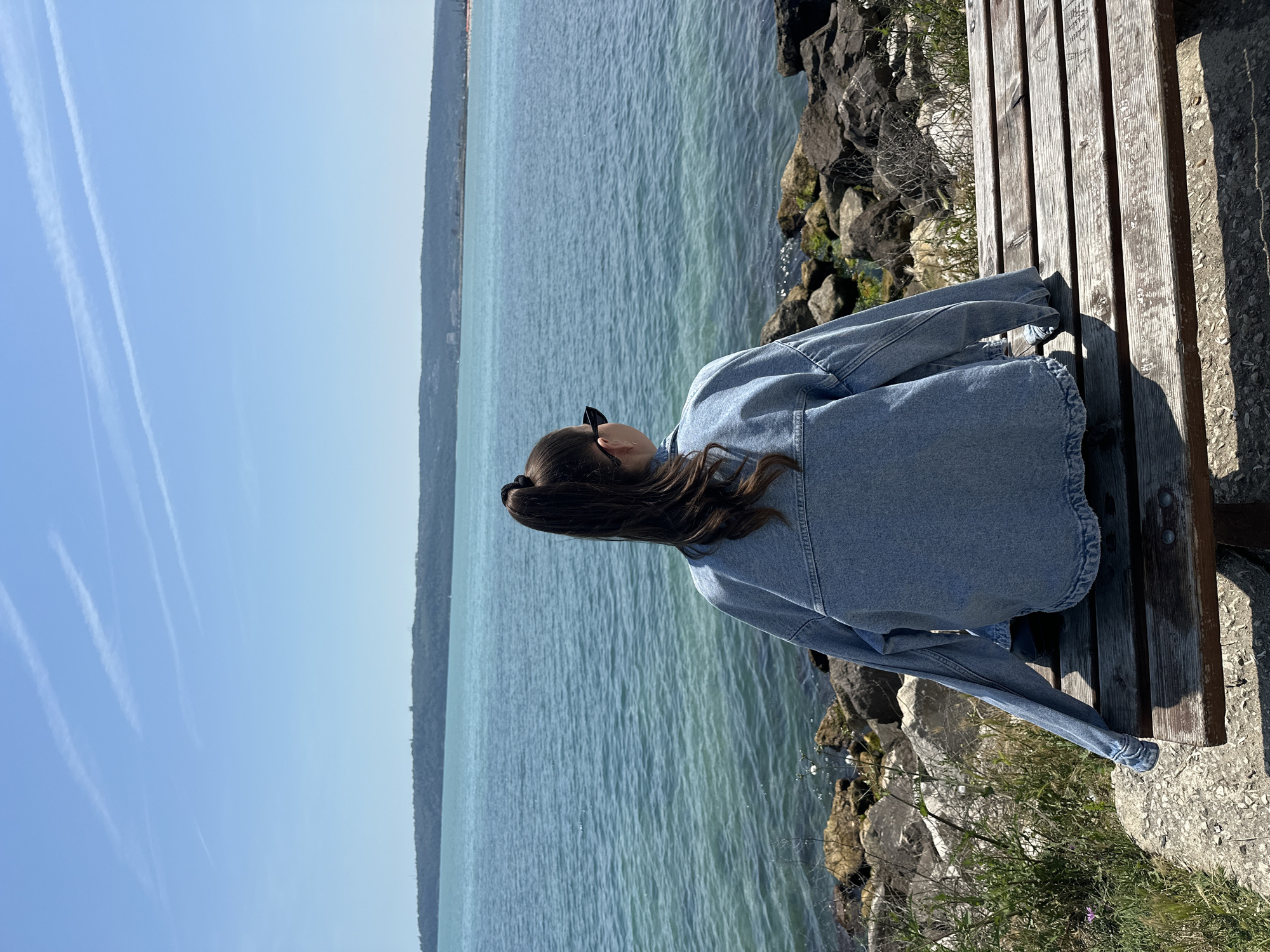 A female individual, viewed from behind, is seated on a weathered wooden bench that runs parallel to a large body of water. She is wearing a light-wash denim jacket, with dark hair pulled into a high ponytail and sunglasses resting on her head. Her left arm, draped over the bench, shows the sleeve extending beyond her hand. The bench sits on a concrete or paved area, immediately bordered by a rocky shoreline composed of various dark and light stones, some with visible algae or moisture. Green vegetation, including grasses and small plants with white blossoms, grows among the rocks and on the adjacent ground. The water, displaying a clear turquoise hue with subtle ripples, extends to a distant, low-lying landmass. This landmass is densely covered in green foliage, with faint indications of structures or buildings along its silhouette. The sky is uniformly light blue and clear, featuring multiple distinct white contrails from aircraft. The scene is brightly illuminated by natural sunlight, casting prominent shadows from the bench and the person. The setting is a coastal or large bay environment in an unspecified city within an unidentified region.