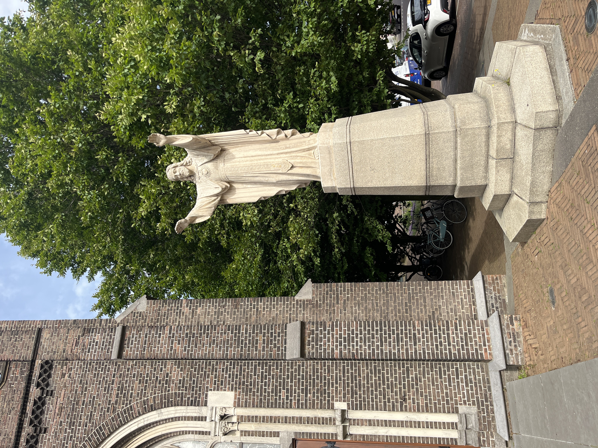 A statue of Jesus Christ stands prominently in front of a brick church in an urban setting. Jesus is depicted with his arms outstretched, wearing a flowing robe. The statue is placed on a multi-tiered stone pedestal. To the left of the statue, a portion of the brick church is visible, featuring an arched window and decorative brickwork. Behind the statue, a lush green tree with dense foliage provides a natural backdrop. In the background, several bicycles are parked near the tree, and a white car is visible on a street alongside other vehicles and traffic signals. The ground is paved with bricks and concrete slabs. The sky above is partially cloudy.