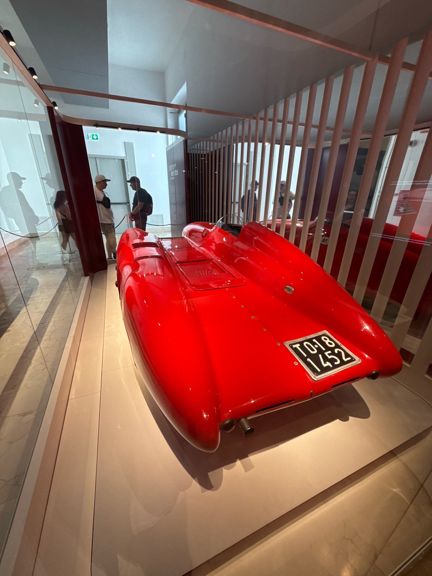 A red vintage sports car is displayed in a museum in Milan, Italy. The car is the focal point, positioned at an angle with its rear facing the viewer. It has a sleek, aerodynamic design and a black license plate with white lettering that reads "TO 18 1452". The car is situated on a raised, light-colored platform, with a polished marble floor reflecting the surroundings.
To the left of the car, a glass partition separates the exhibit from a walkway where several people are visible. Two men are standing and talking, one wearing a white cap and the other a black cap. Further left, a woman and another person are silhouetted against a white background.
Behind the car, vertical wooden slats create a decorative screen, hinting at the museum's architecture. Through the slats, another red object, possibly another car or part of an exhibit, can be seen. The ceiling is modern, with track lighting. The environment appears to be a well-lit, contemporary exhibition space designed to showcase historical artifacts.