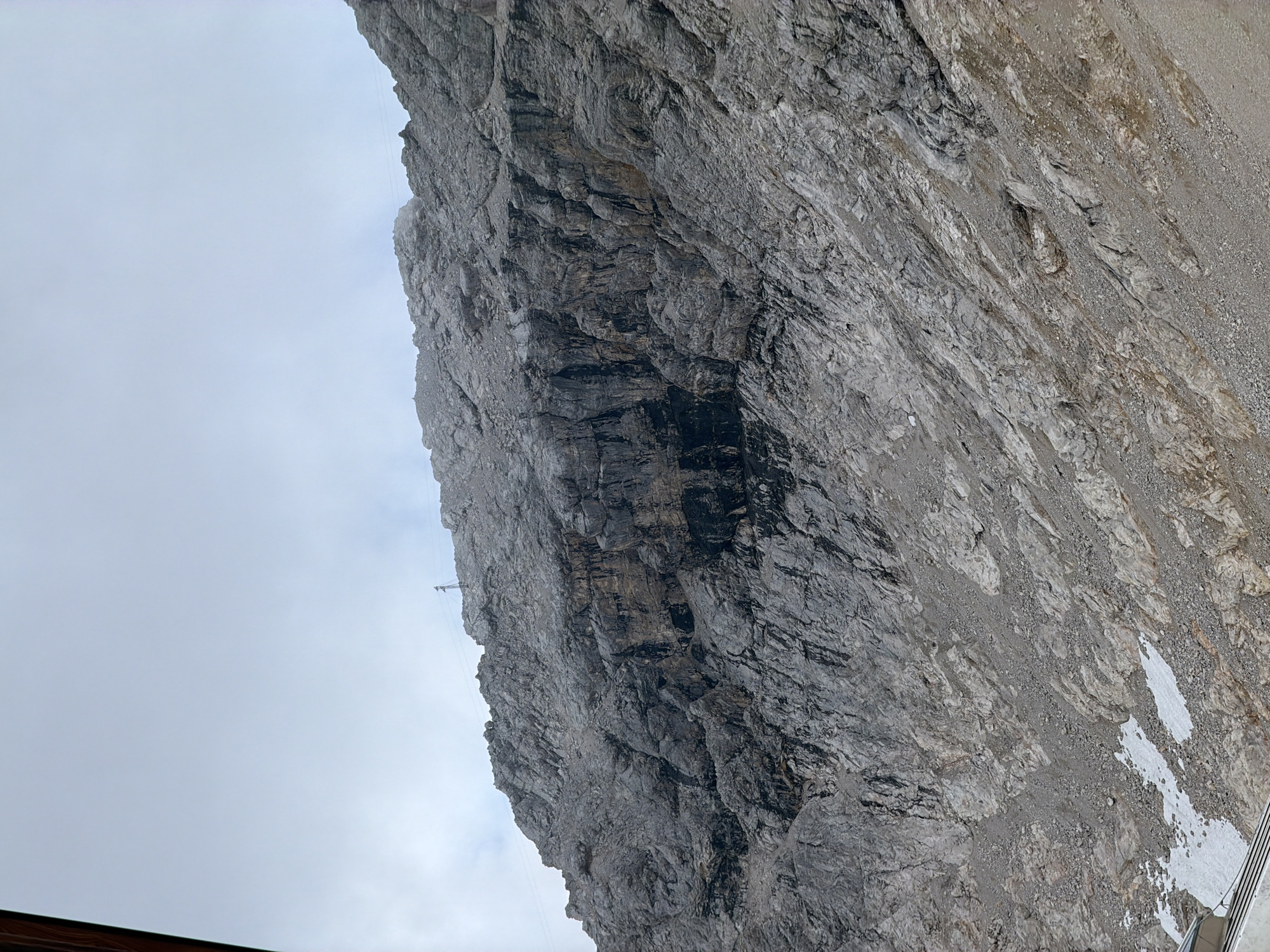 The image depicts a rocky, mountainous terrain under a cloudy sky. The mountain face is steep and covered in various sizes of rocks and gravel. There are patches of snow visible towards the bottom left of the image. A small section of what appears to be a metal structure or railing is visible in the bottom left corner. A solitary transmission tower or antenna is visible on the peak of the mountain in the upper right portion of the image. The scene is devoid of any visible people or animals. This landscape is characteristic of the Zugspitze, Germany's highest peak, located near Garmisch-Partenkirchen.