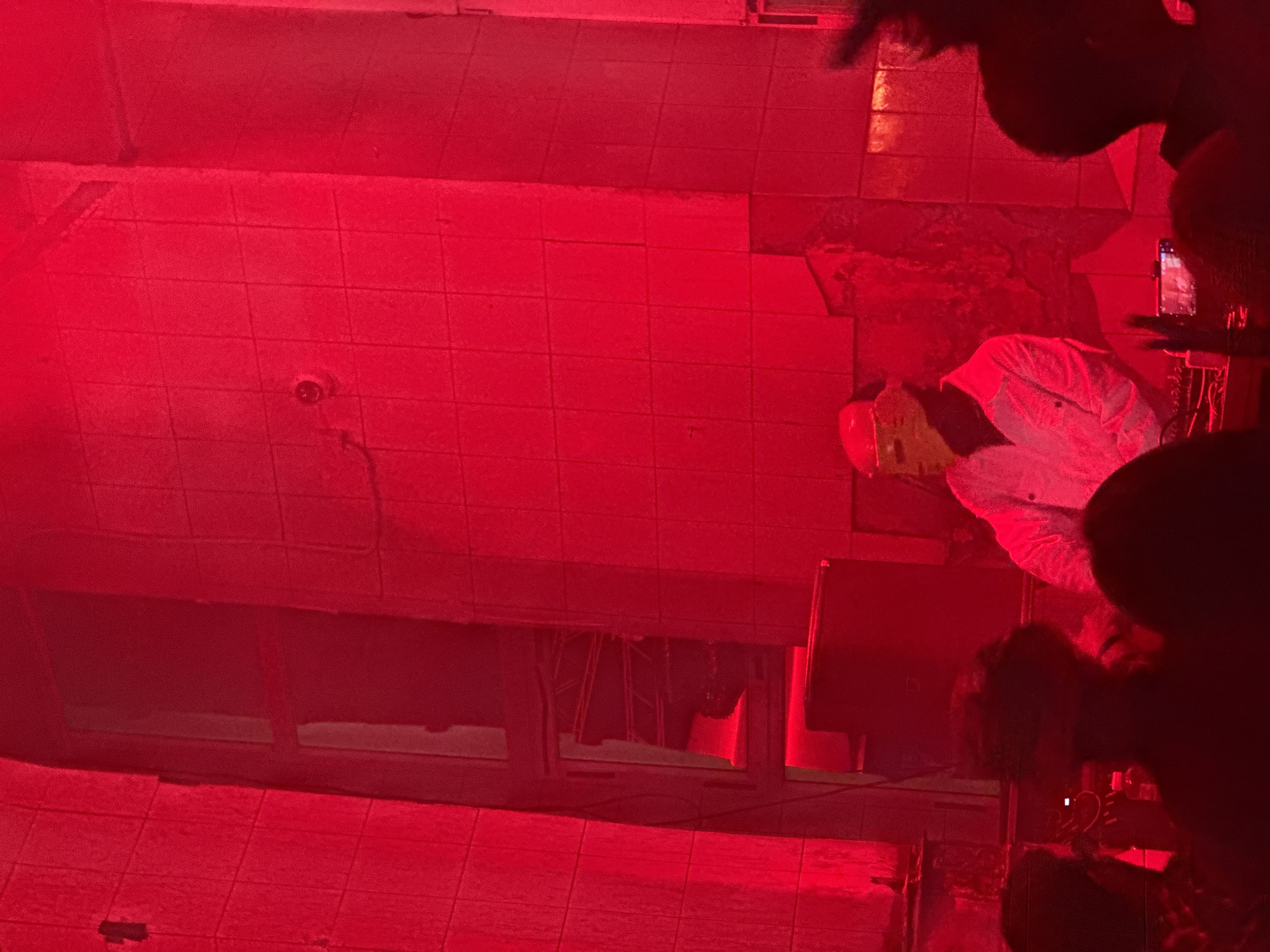 The scene depicts an indoor event in Moscow, Russia, illuminated entirely by a deep red light source. Centrally positioned behind a console, a performer stands, wearing a light-colored long-sleeved shirt. This individual's face is obscured by a reflective visor featuring a prominent horizontal band across the eye level. To the performer's immediate left, a large, dark speaker or monitor is situated. The background consists of light-colored tiled walls, some sections exhibiting visible damage, including cracks and missing tiles, exposing underlying darker material. Vertical structural divisions or pillars frame the central area, revealing darker, recessed openings with indistinct internal structures. In the immediate foreground, the dark silhouettes of an audience are visible, with one person on the right holding a smartphone vertically, its screen faintly illuminated.