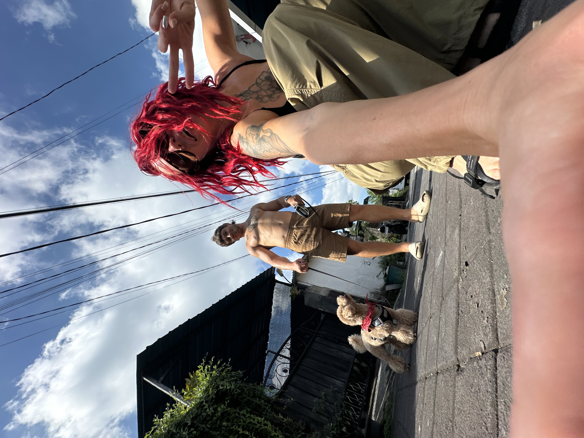 Captured from a low angle, the image shows a woman with red hair giving the peace sign. She is wearing sunglasses and has tattoos on her chest and arm. Above her, an arm reaches into the frame. The woman sits next to a person in khaki pants. In the background, a shirtless man walks a dog on a leash. The environment includes a roof, plants, a fence, power lines against a partly cloudy sky, and a paved road. The scene suggests a location in the city of North Kuta, Indonesia.