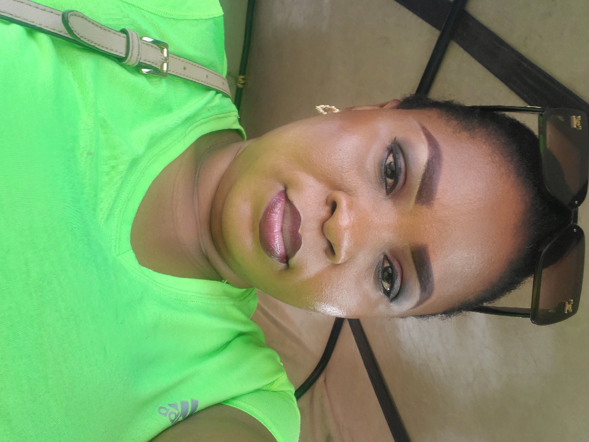 A portrait of a Black woman, with the image rotated 90 degrees clockwise, so her head is tilted to the right. She looks directly at the camera with a gentle, pleasant smile.
She is wearing a bright, neon green short-sleeved top or t-shirt. A gray Adidas logo, depicting three stripes in a triangular arrangement, is visible on the lower left side of her shirt. A light beige strap, likely from a bag, with a gold-toned buckle, crosses her upper chest.
Her dark hair is styled back. She has distinct, shaped eyebrows and dark eye makeup, including eyeliner and eyeshadow, enhancing her eyes. Her lips are covered in a glossy pink or mauvy lipstick. A small, gold-colored earring is visible on her left ear. A pair of dark sunglasses, featuring a designer-style logo (resembling interlocking letters such as 'LV' or a similar monogram) on the temple, is perched on top of her head.
Her skin appears smooth and has a dewy sheen. The background is out of focus, showing indistinct light brown and beige tones, along with a dark strap or bar, suggesting an indoor or shaded outdoor setting. The scene conveys a casual atmosphere in Lagos, Nigeria.