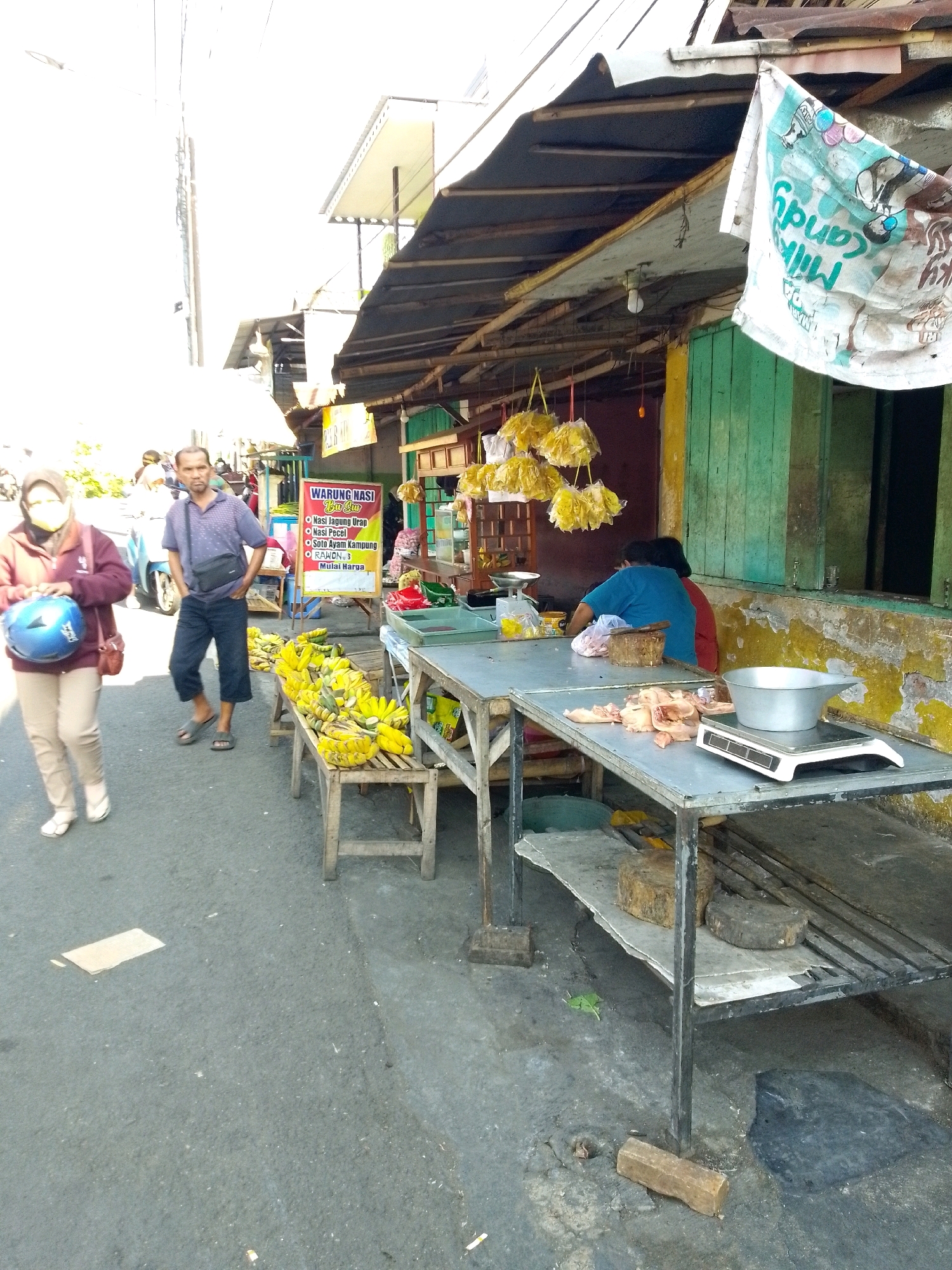 The image depicts a street-level view of a market stall in Gebang, Indonesia. The stall appears to be selling food items, with a prominent display of bananas on a wooden stand in the foreground. Various other items, including raw chicken pieces, are arranged on metal tables, some of which are equipped with scales. A banner advertising "WARUNG NASI BU LULUK" (Rice Stall Bu Luluk) is visible, listing menu items such as "Nasi Jagung Urap," "Nasi Pocel," and "Soto Ayam Kampung."
In the mid-ground, a man in a blue polo shirt and dark pants is standing near the banana display, looking towards the camera. To his left, a woman wearing a maroon jacket and beige pants, with a blue helmet in her hand, walks away from the camera. A motorcycle is parked behind her.
The stall itself is covered by a weathered awning, and the building behind it has green wooden shutters. In the background, other stalls and activity suggest a bustling marketplace. The overall environment is an outdoor urban setting with paved ground and a bright, sunny atmosphere.