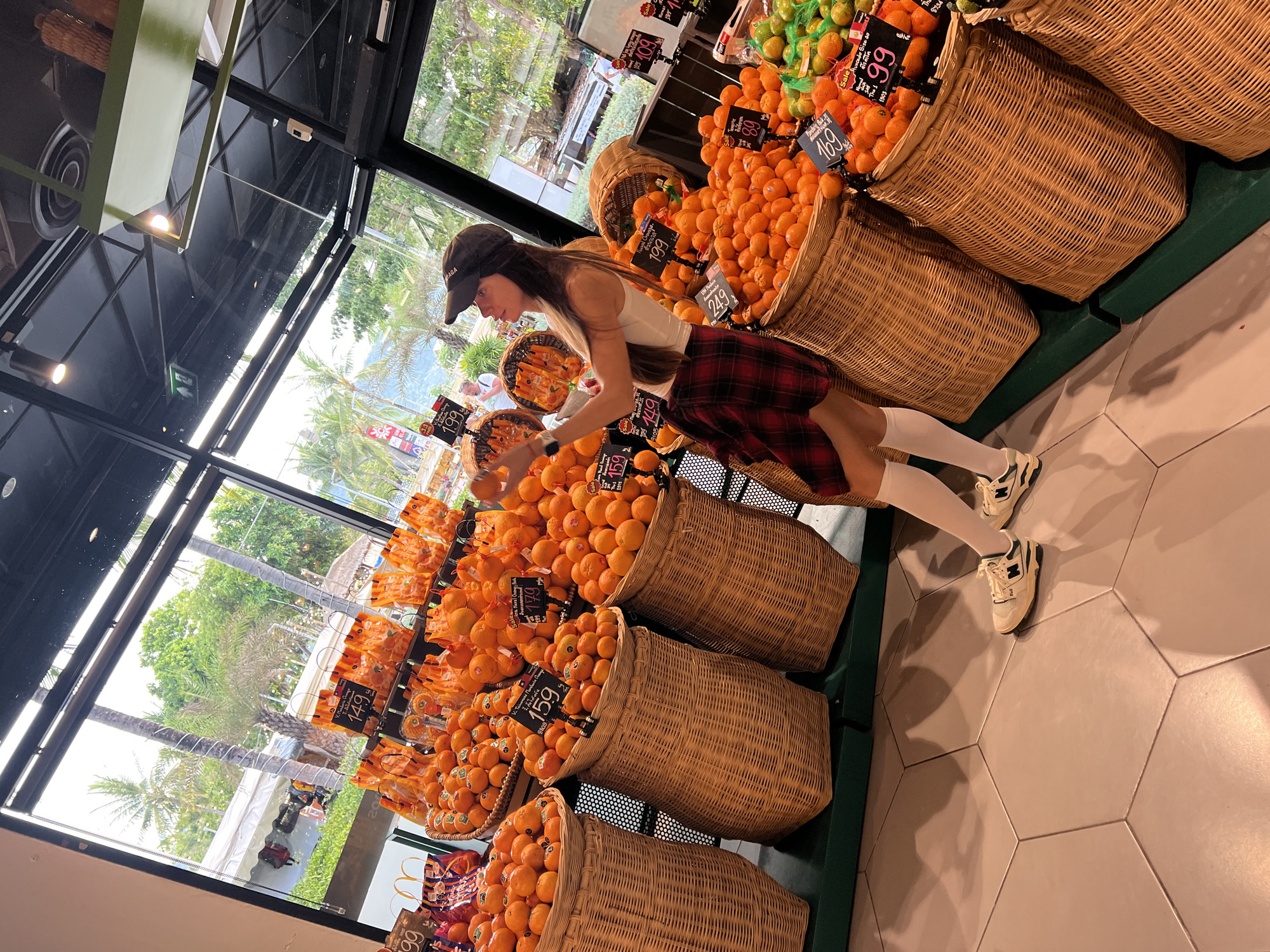 The image shows a woman shopping for oranges in what appears to be a grocery store in Baan Chaweng, Thailand. The woman, who is wearing a white tank top, a red plaid skirt, white knee-high socks, and white sneakers, is standing next to several large wicker baskets filled with oranges. The baskets have price tags on them that include numbers like "149," "159," "169," and "199." A window to the left of the scene shows palm trees and other tropical vegetation, and the sky is visible in the background. The store itself has a modern interior with a black ceiling, and there are other baskets of oranges visible in the background.