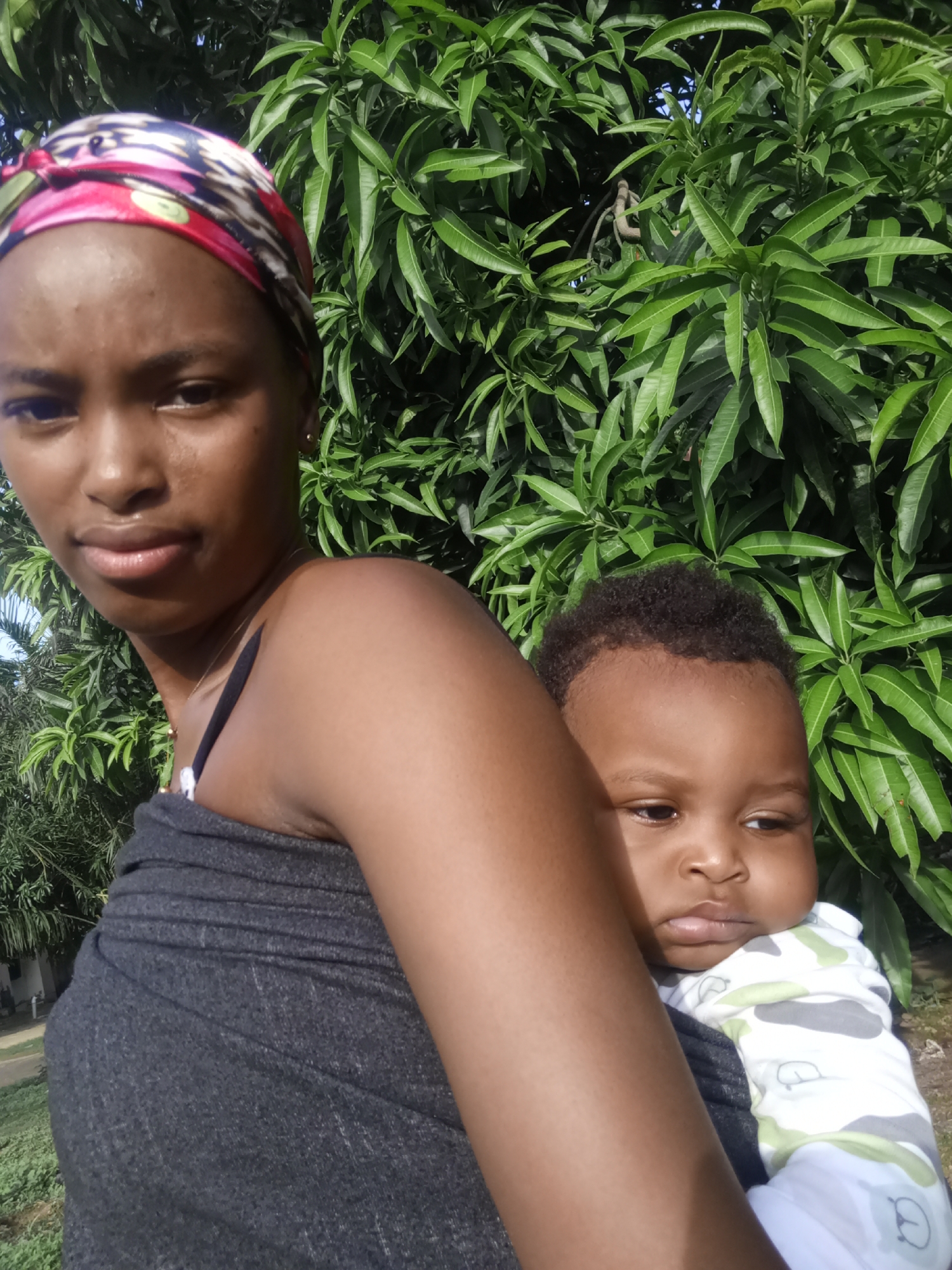 A young woman is holding a baby against a backdrop of lush foliage, possibly in the city of Keffi, Nigeria. The woman is dark-skinned and has a red, black, and white headscarf. She is wearing a gray wrap-like garment over a black tank top, with a thin gold necklace visible. The baby is also dark-skinned, and is wearing a light-colored onesie. The background consists primarily of a leafy green tree. The photo appears to be taken outdoors.