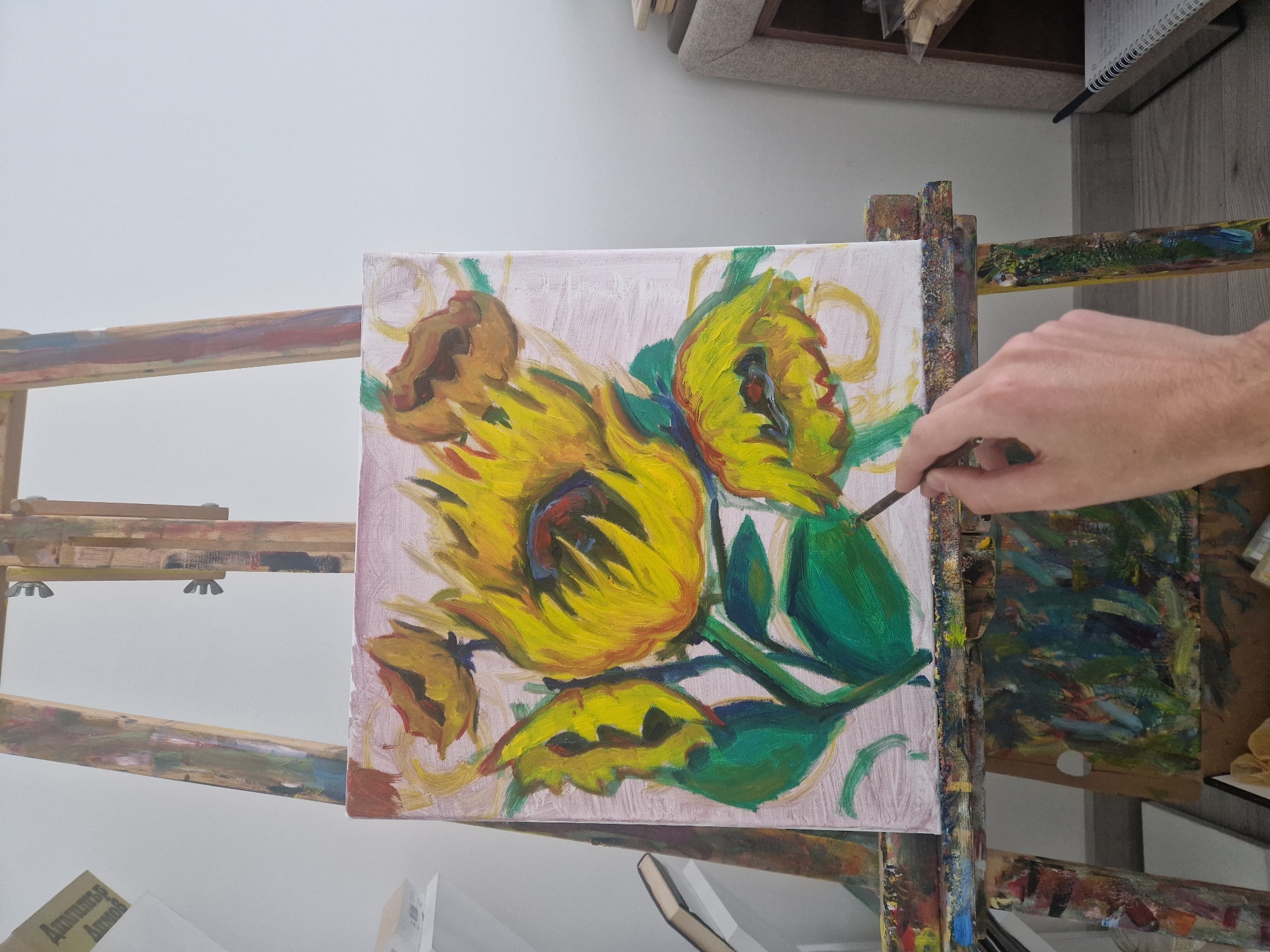 A partially completed oil painting of sunflowers is displayed on a wooden easel. The artist's hand, holding a brush, is visible to the right, suggesting the painting is in progress. The painting features bold brushstrokes and vibrant colors, primarily yellows and greens, against a pale background. The scene is set indoors, likely in an artist's studio, indicated by the presence of another easel and art supplies in the background.