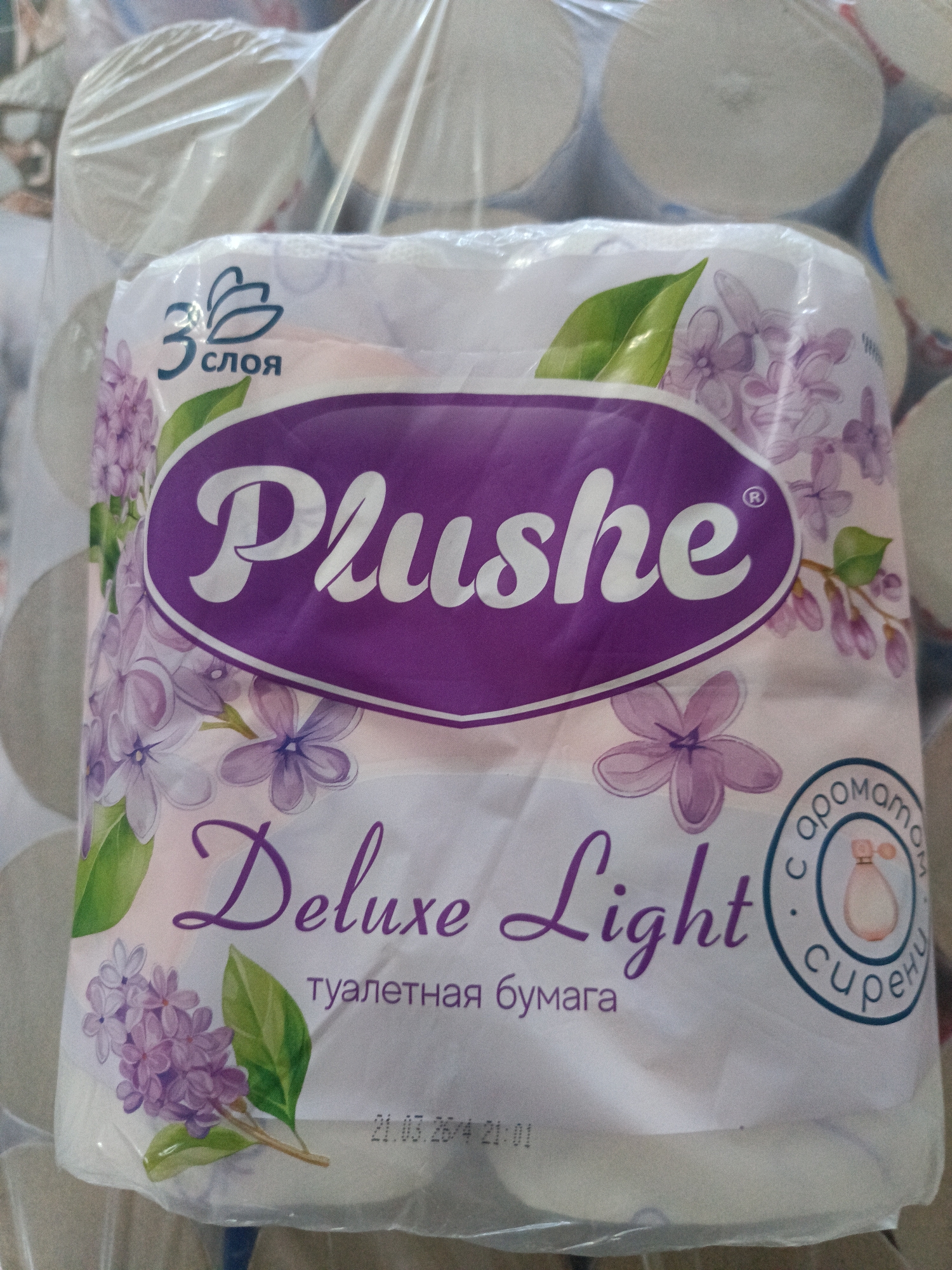 This media file shows a close-up of a package of "Plushе" brand toilet paper. The packaging is predominantly white and purple, adorned with illustrations of lilac flowers and green leaves. The brand name "Plushе" is prominently displayed in large, stylized white lettering within a purple oval. Below the brand name, the words "Deluxe Light" are written in a decorative purple script, followed by the Russian text "туалетная бумага" (toilet paper).
In the upper left corner, there is a logo indicating "3 слоя" (3 layers) above a stylized graphic. To the right, a circular emblem with blue text and a pink perfume bottle illustration is visible, with Russian text that appears to read "с ароматом сирени" (with lilac scent). A date and time stamp, "21.03.24 21:01," is printed near the bottom. The background suggests the package is part of a larger stack of toilet paper rolls, wrapped in clear plastic. The lighting is bright and appears to be artificial, as there are no visible shadows suggesting natural light sources. The scene is an indoor product shot, likely in a retail or storage environment. No people are visible. The focus is solely on the product packaging.