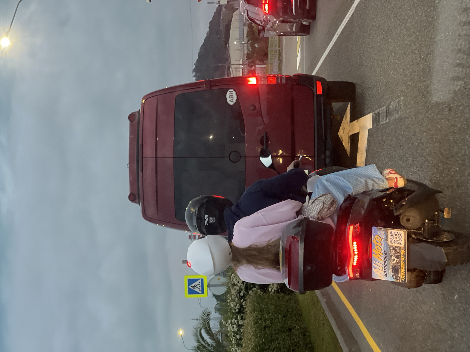 A two-person moped, with both riders wearing helmets, is behind a red van on a paved road in Sochi, Russia. The rear of the van is visible, including the license plate area, with an "ABH" sticker. The moped's license plate is visible and includes Russian text "МОТОПРОКАТ" and a phone number "+7(930)807-47-50". The male driver is wearing a black helmet, and the female passenger is wearing a white helmet and a light pink top. The sky is overcast. A yellow pedestrian crossing sign is visible on the left side of the road, along with some greenery. A streetlight is also visible in the background. In the distance, a building or structure is visible on a hillside. An arrow indicating the direction of travel is painted on the road.