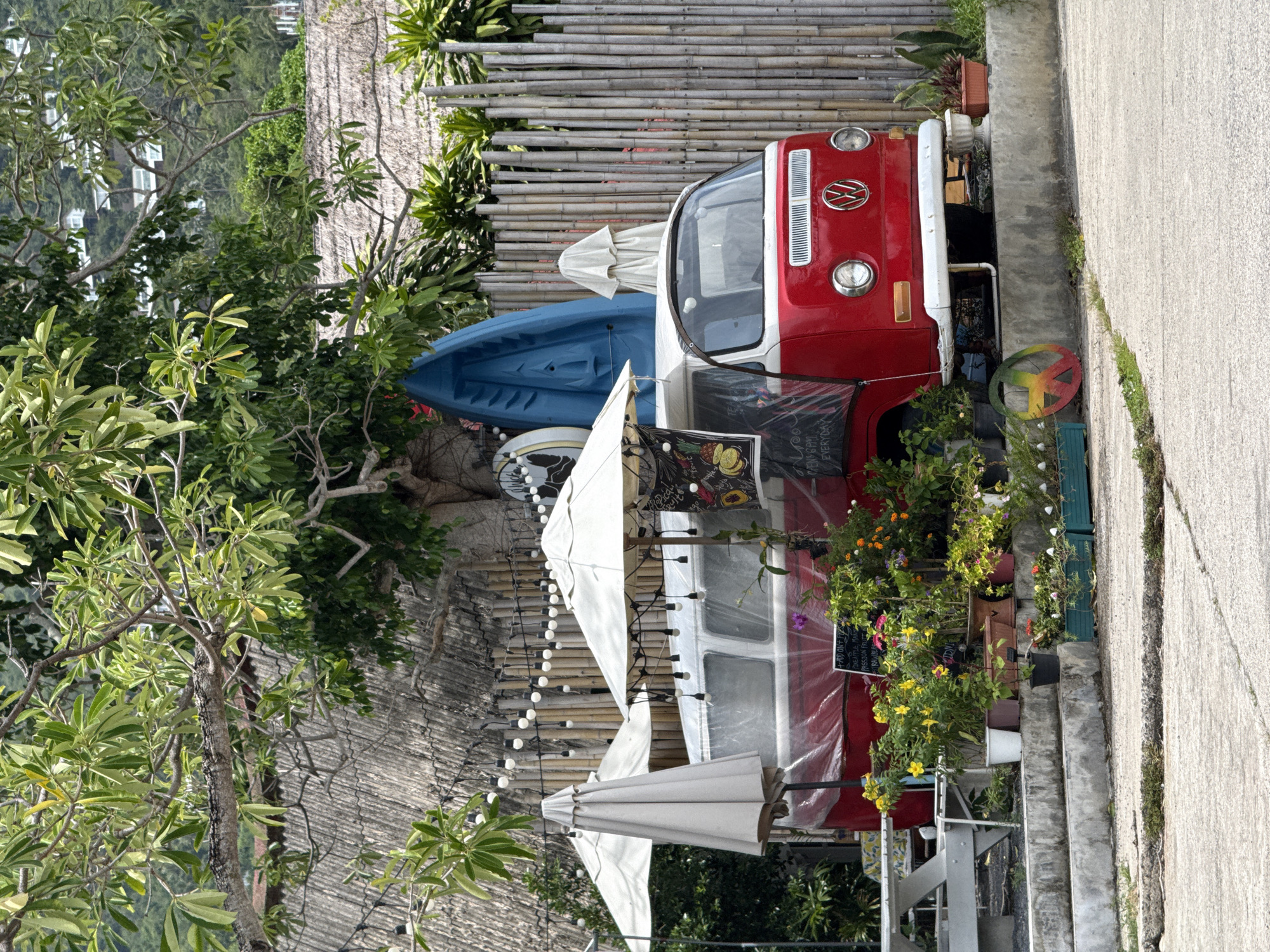 This image depicts an outdoor scene in Ko Samui District, Thailand, featuring a red and white Volkswagen Type 2 Kombi van serving as a food or drink stand. The van is parked on a concrete surface, facing forward. Behind the van, a tall bamboo fence with a woven thatched roof structure above it runs horizontally. Various potted plants, including flowering bushes with yellow and pink blossoms, are arranged in front of and around the van. Two white umbrellas are set up near the van, providing shade. A blue kayak or surfboard is leaning against the back of the van. String lights are visible hanging along the bamboo fence. The background shows lush greenery and a hillside with buildings. A picnic table with benches is positioned to the left of the van. The overall impression is a casual, tropical setting.