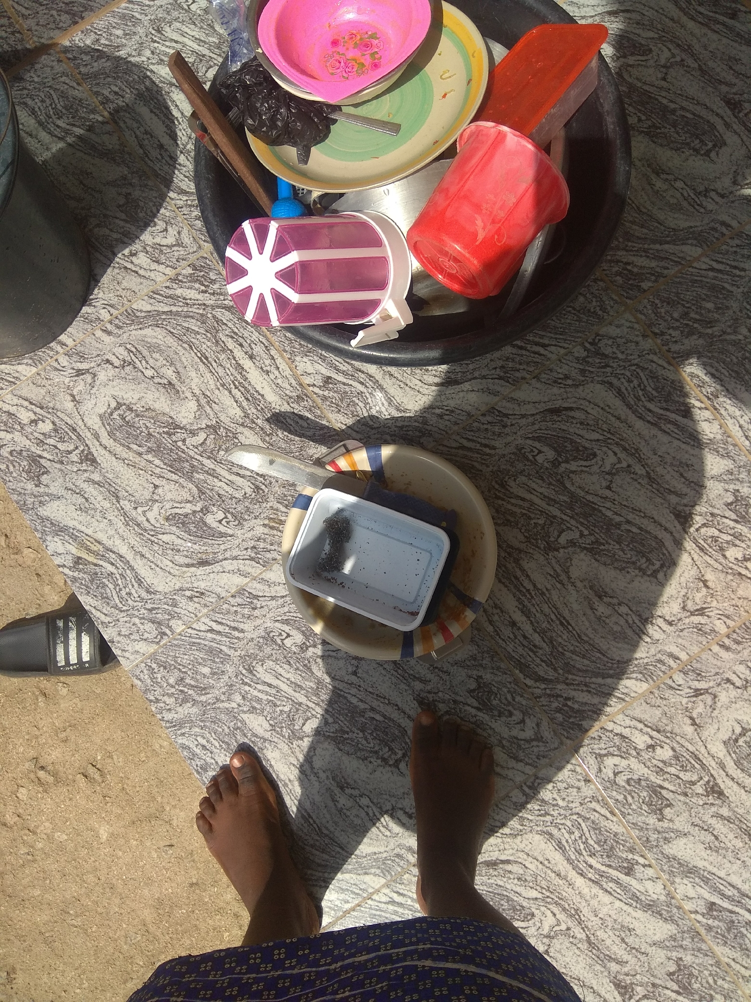 The overhead shot captures a scene of washing dishes outside on a sunny day. Several plastic containers and plates are piled in a large black basin. Among them are a pink bowl decorated with flowers, a red plastic cup, and a white plastic container with a purple lid. A metal utensil rests on the edge of the basin. A pair of dark-skinned feet and part of a patterned blue skirt are visible at the bottom of the frame, suggesting someone is standing over the washing station. A black flip-flop lies to the left. The ground is tiled with a swirling gray and white pattern, interspersed with sandy areas. The strong shadows indicate bright sunlight, likely midday. The scene is set outdoors, possibly in a yard or patio area.