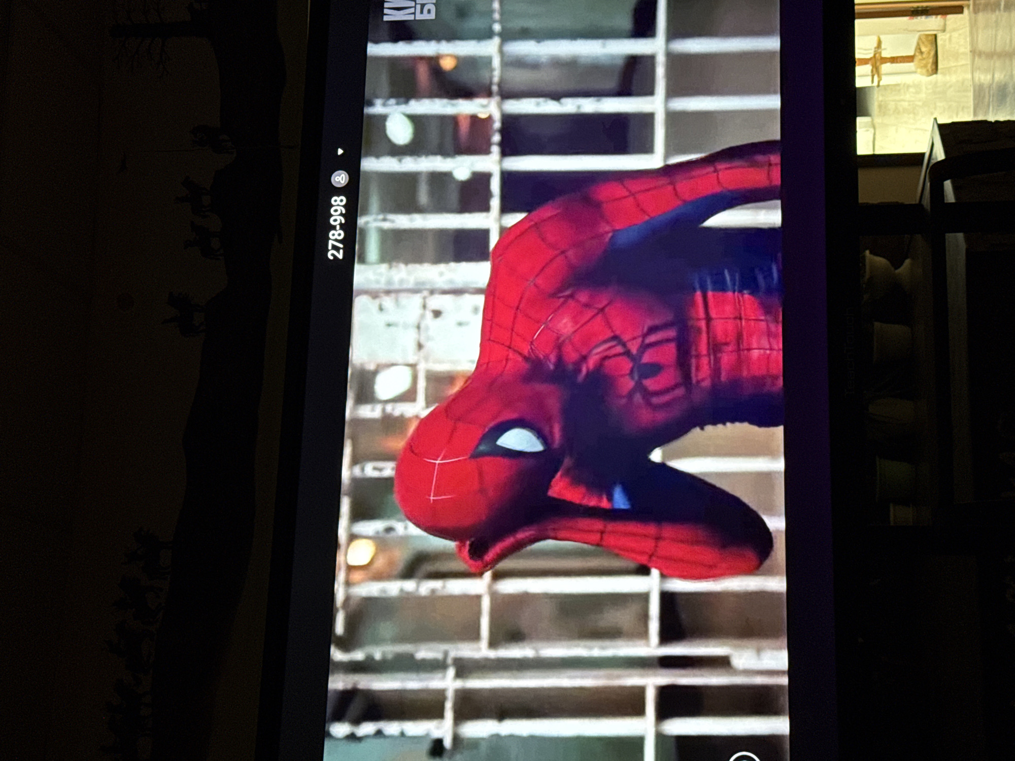 The image displays a television screen showing a scene with the character Spider-Man in a red and blue suit. He is positioned in what appears to be a jail cell, with a bar-like structure behind him. His left hand is raised to his forehead, suggesting a gesture of distress or contemplation. The top of the screen shows a banner with "278-998" and a user icon. The background above the screen shows a silhouetted decorative frieze depicting horses and riders, possibly referencing a historical or western theme. The scene takes place indoors, with the TV screen reflecting ambient light. There is no specific indication of the location being Nyurba, Russia.