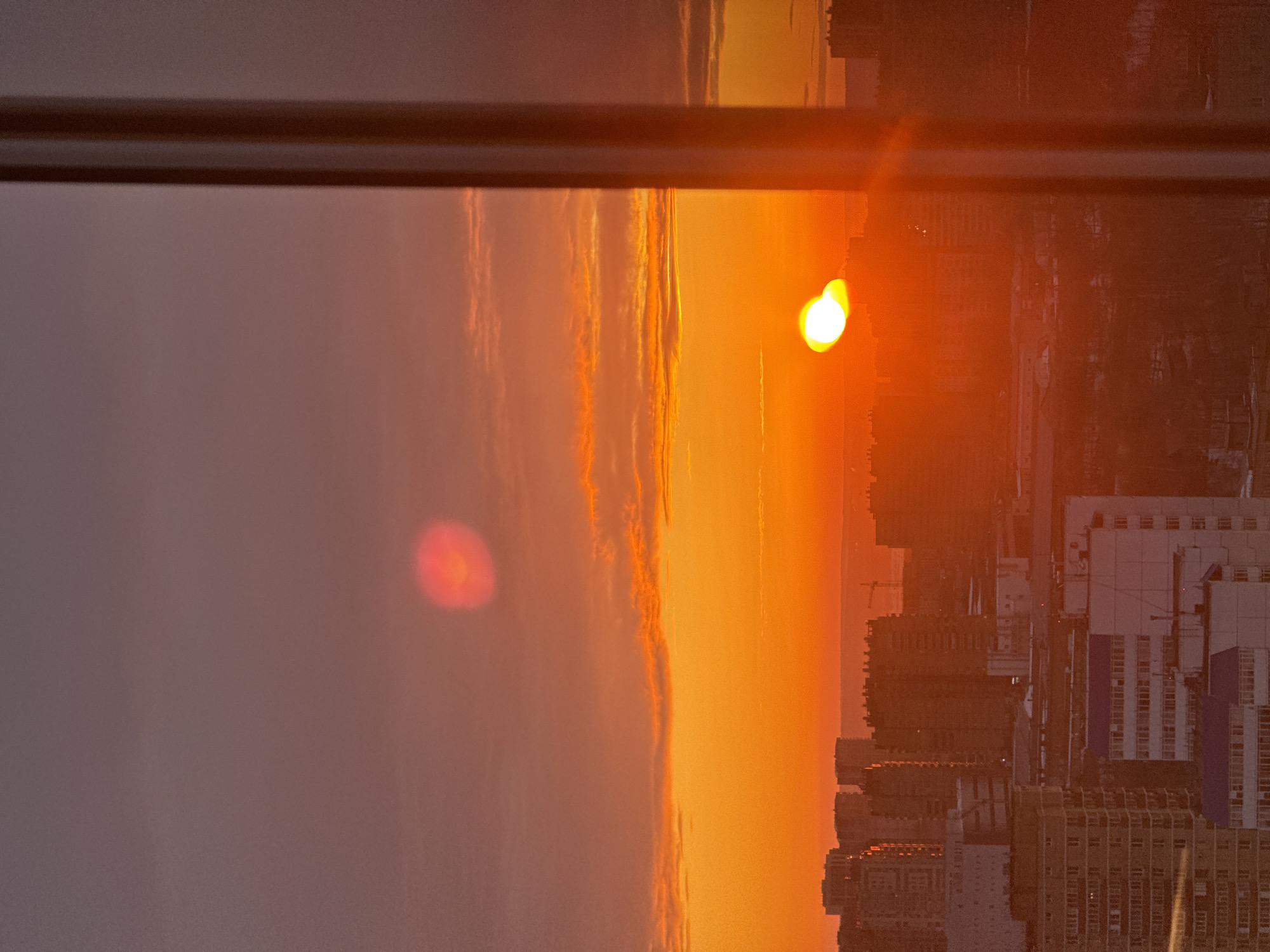 The image depicts an elevated view of the city of Perm, Russia, during either sunrise or sunset. The sky exhibits an intense orange-red glow along the horizon, transitioning to a deeper reddish-purple in the upper segments, featuring elongated cloud formations illuminated by the sun. The sun appears as a bright, distorted yellow-white sphere positioned near the center-right, partially obscured by the city skyline. Prominent lens flare is present, including a circular red-orange anomaly in the upper-left sky and a vertical light streak along the right side of the frame. The cityscape consists of numerous multi-story residential and commercial buildings, predominantly silhouetted against the bright horizon. In the lower-middle foreground, some buildings exhibit white facades and blue roofs. A distant horizon line reveals faint topographical features and a silhouetted construction crane. A dark, vegetated area is visible interspersed among the structures. A dark, vertical structural element, consistent with a window frame or pillar, occupies the rightmost portion of the image. No individuals or specific actions are discernible.